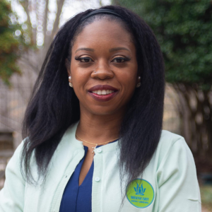 A woman outdoors wearing a light green jacket with a green and blue badge, smiling at the camera.