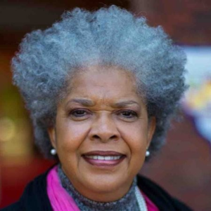 Close-up of an elderly woman with gray curly hair, wearing a pink and black top, smiling outdoors.