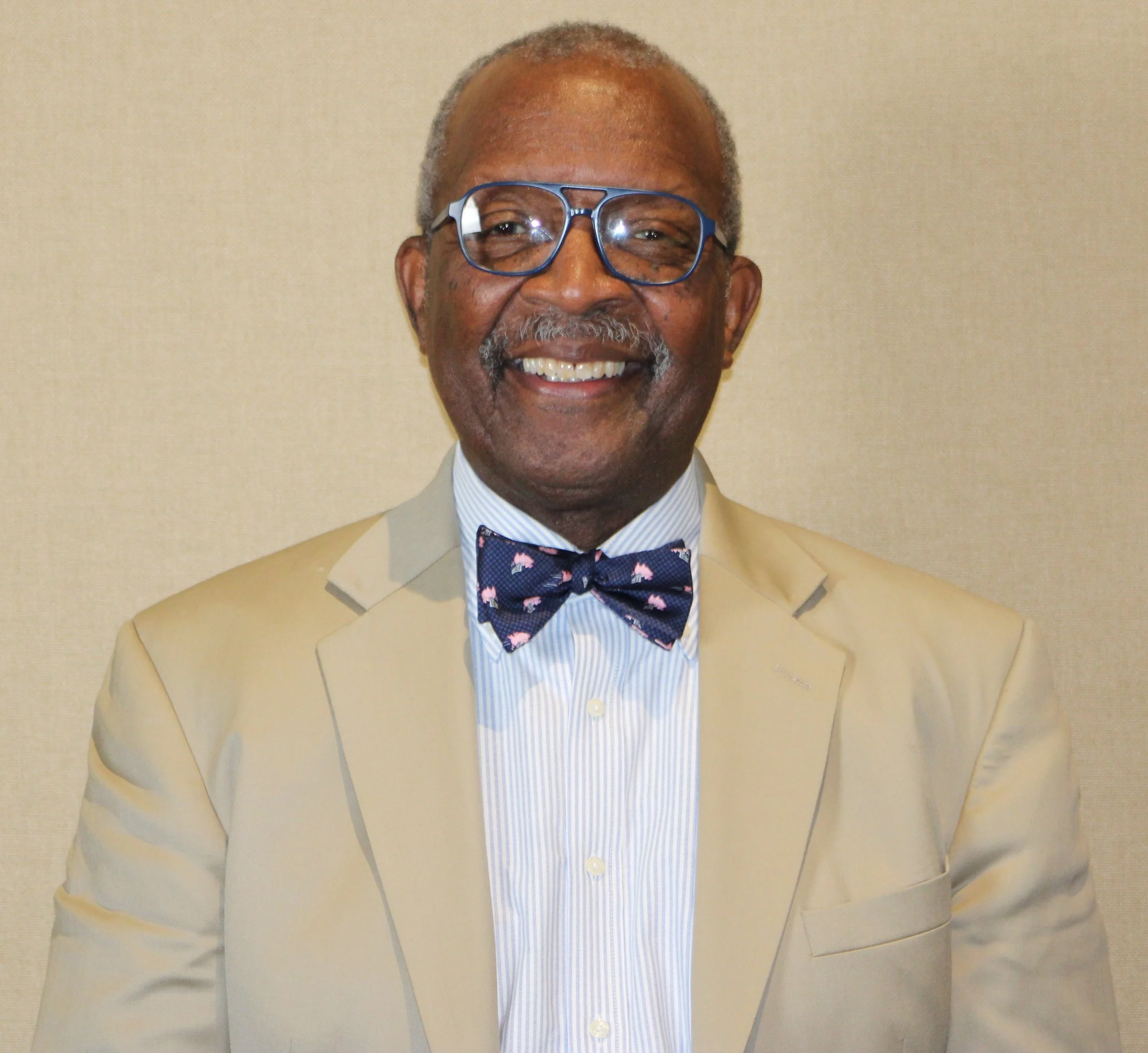 A smiling older African American man wearing a beige suit, light blue striped dress shirt, navy blue bow tie with pink patterns, and glasses, standing against a plain beige wall.