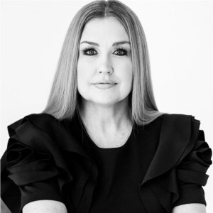 A black and white portrait of a woman with long straight hair, wearing a dark top with ruffled sleeves, looking at the camera with a slight smile.