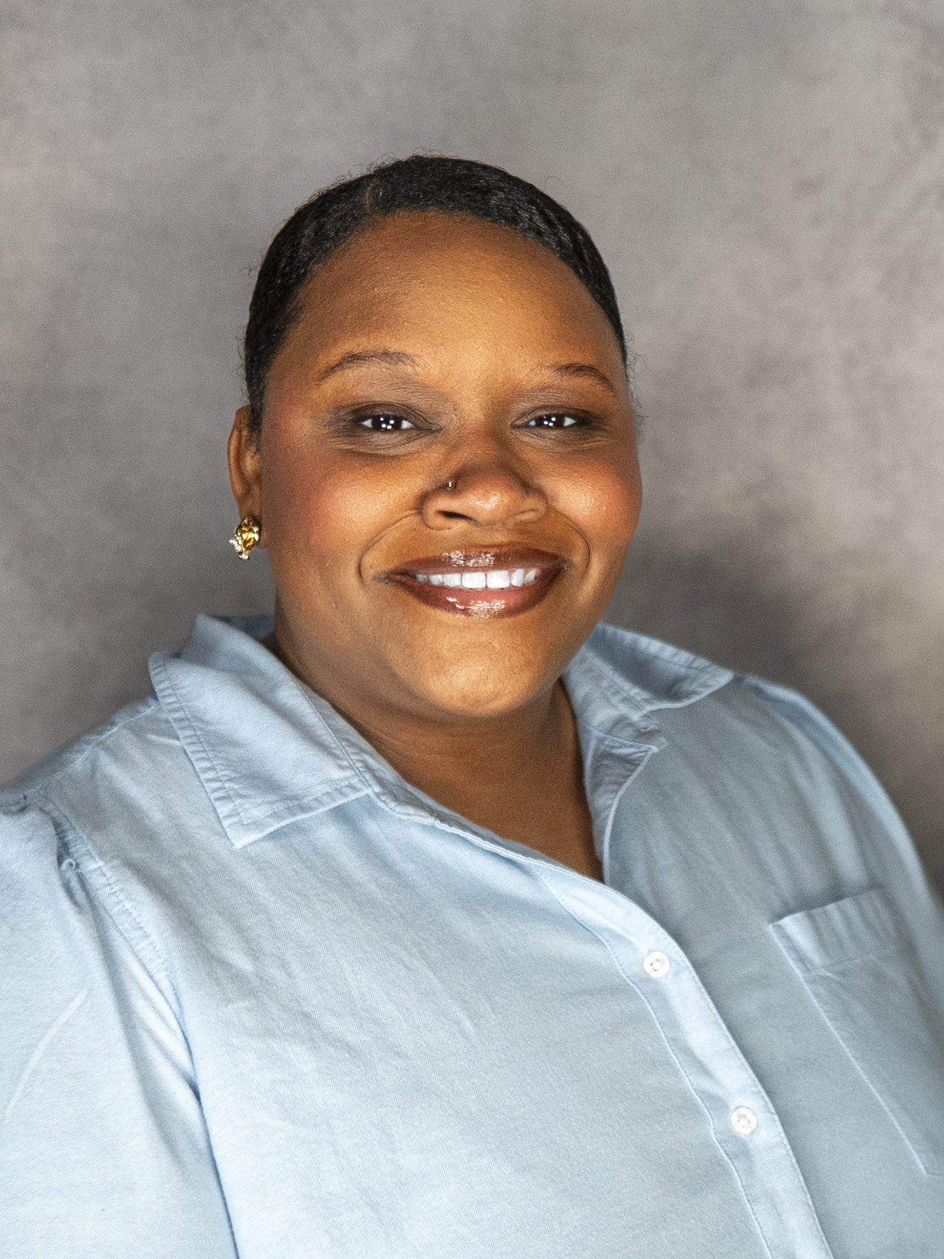 A smiling woman with short hair, wearing a light blue button-up shirt and gold earrings, posing against a neutral background.