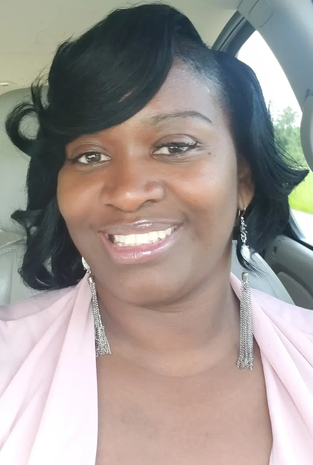 A woman with black, wavy hair, smiling, wearing dangling earrings and a light pink blazer, sitting in a car with a green landscape outside the window.