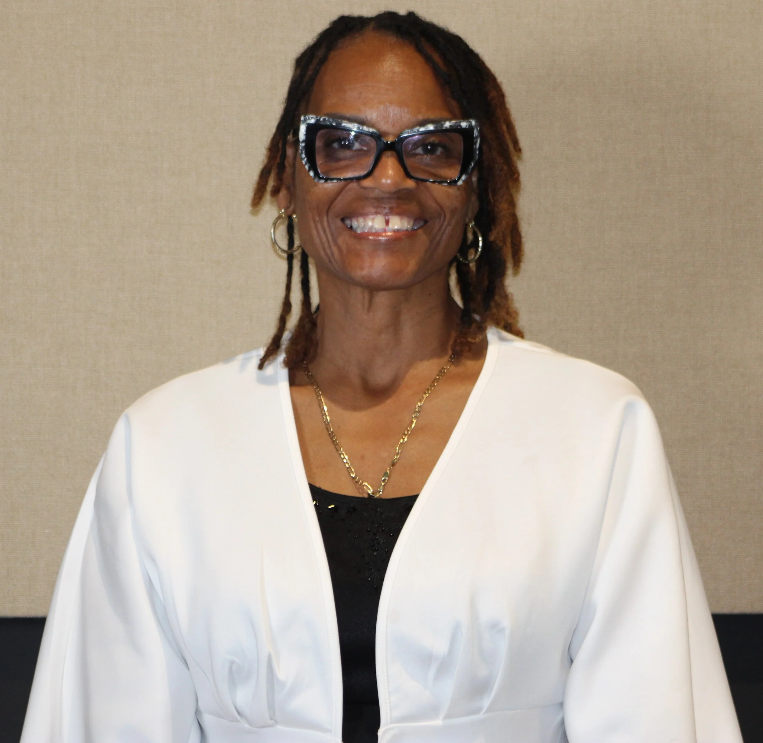 Portrait of a smiling African American woman with glasses, wearing a white blazer and gold jewelry, standing against a beige background.