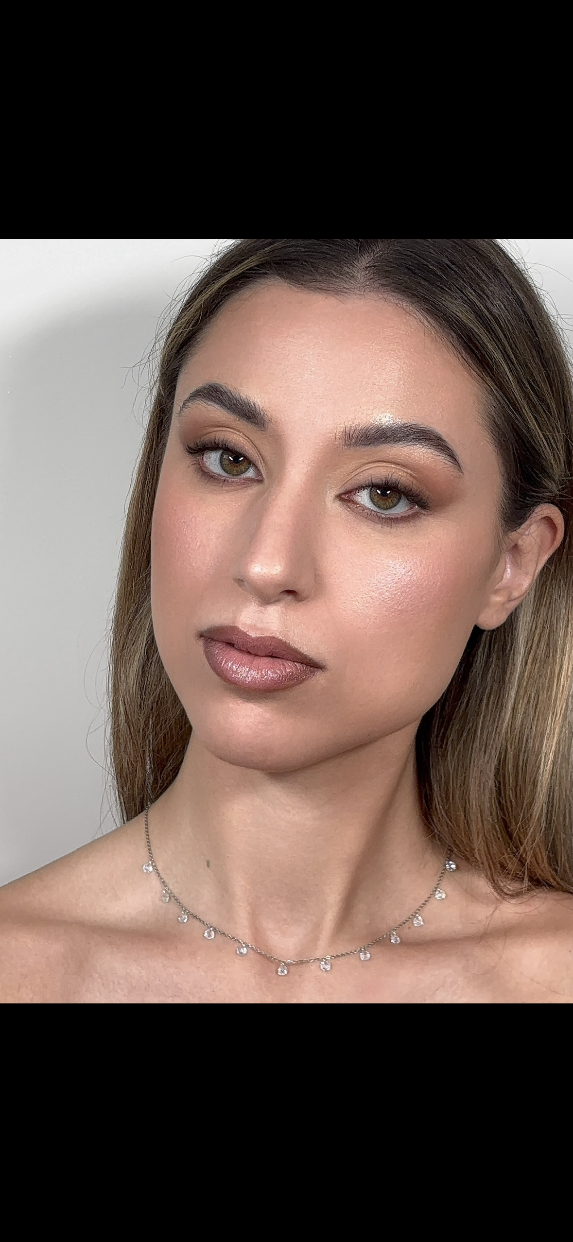 Close-up portrait of a woman with light skin, brown hair, hazel eyes, wearing makeup and a jewelry necklace with small pendants, against a plain white background.