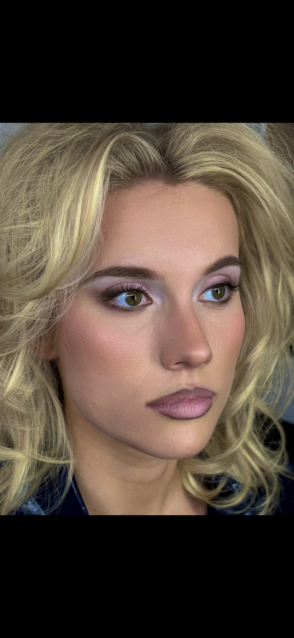 Close-up of a woman with blonde, styled hair, wearing makeup with light eyeshadow, eyeliner, and soft pink lipstick.