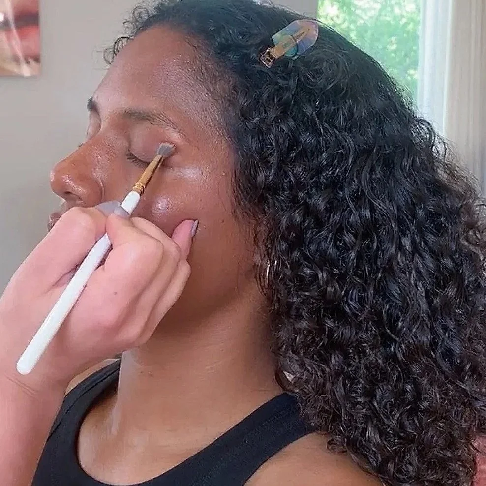 A woman with dark curly hair is getting her eyeshadow applied by a makeup artist using a brush, with a clip in her hair and a window in the background.