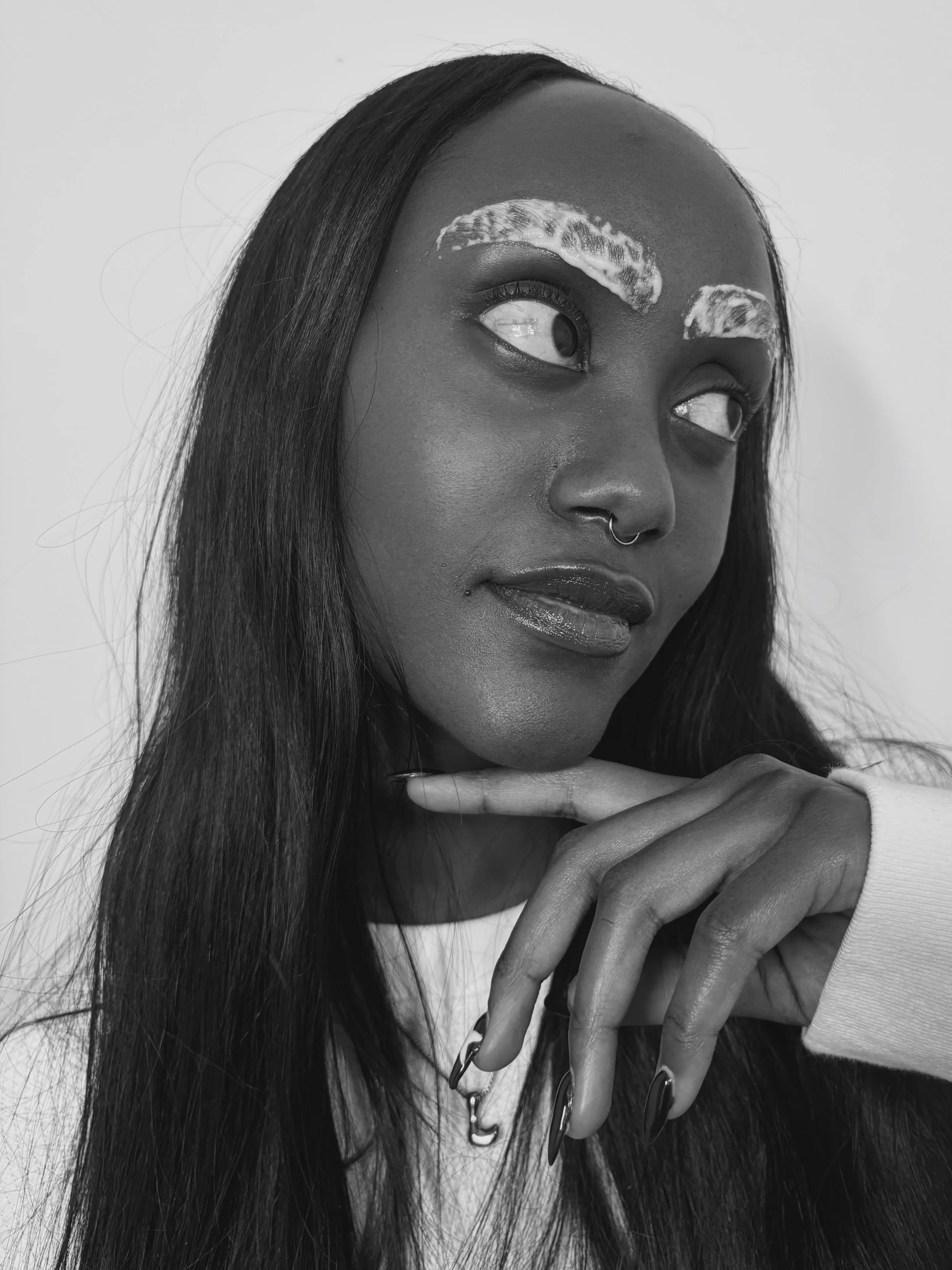 Black and white portrait of a woman with long hair, wearing a septum ring, with painted eyebrows, looking off to the side, resting her chin on her hand.