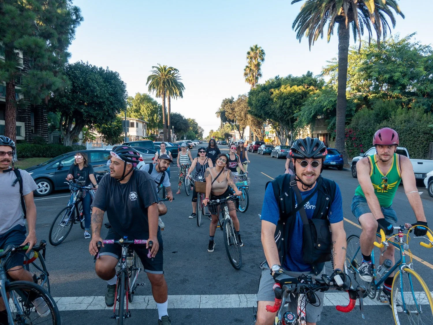 Group of people riding bicycles on a city street with trees and parked cars.