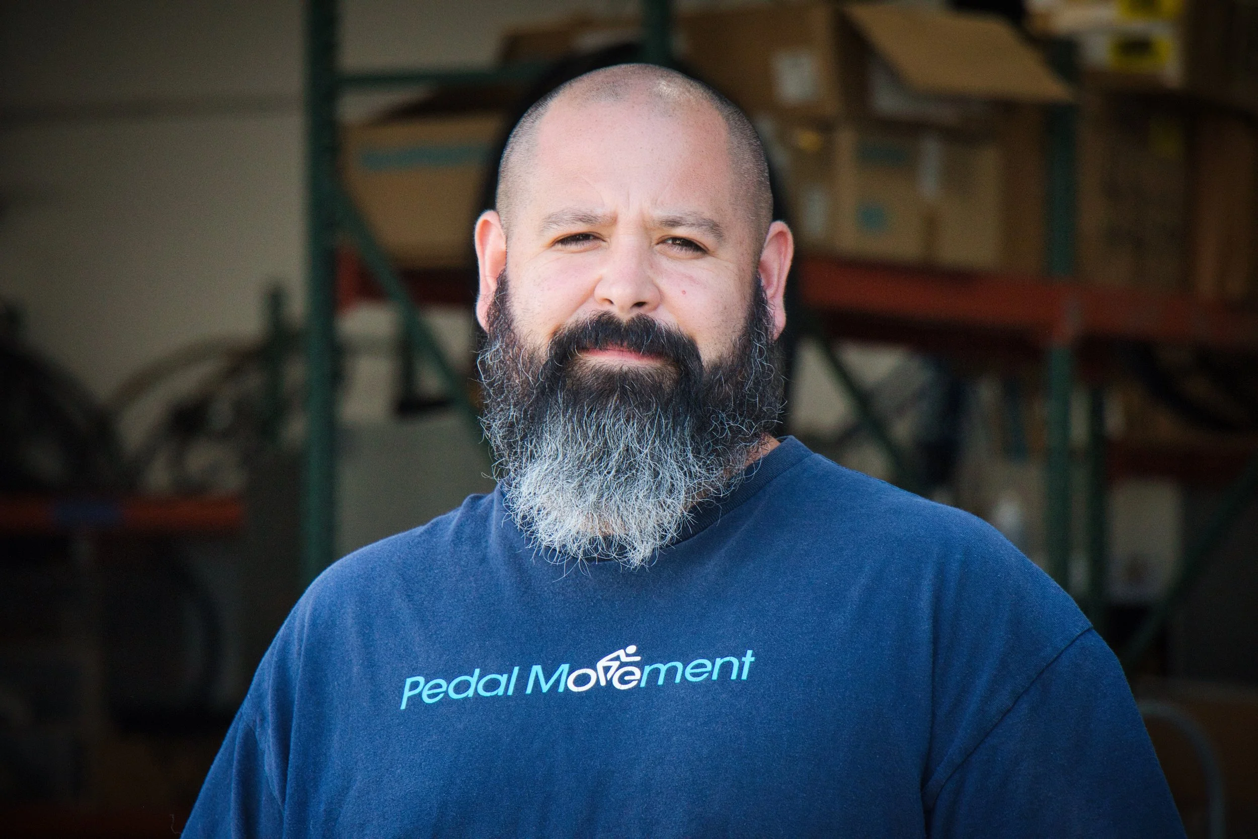 A man with a shaved head, beard, wearing a blue Pedal Movement T-shirt, standing in a workshop or warehouse.