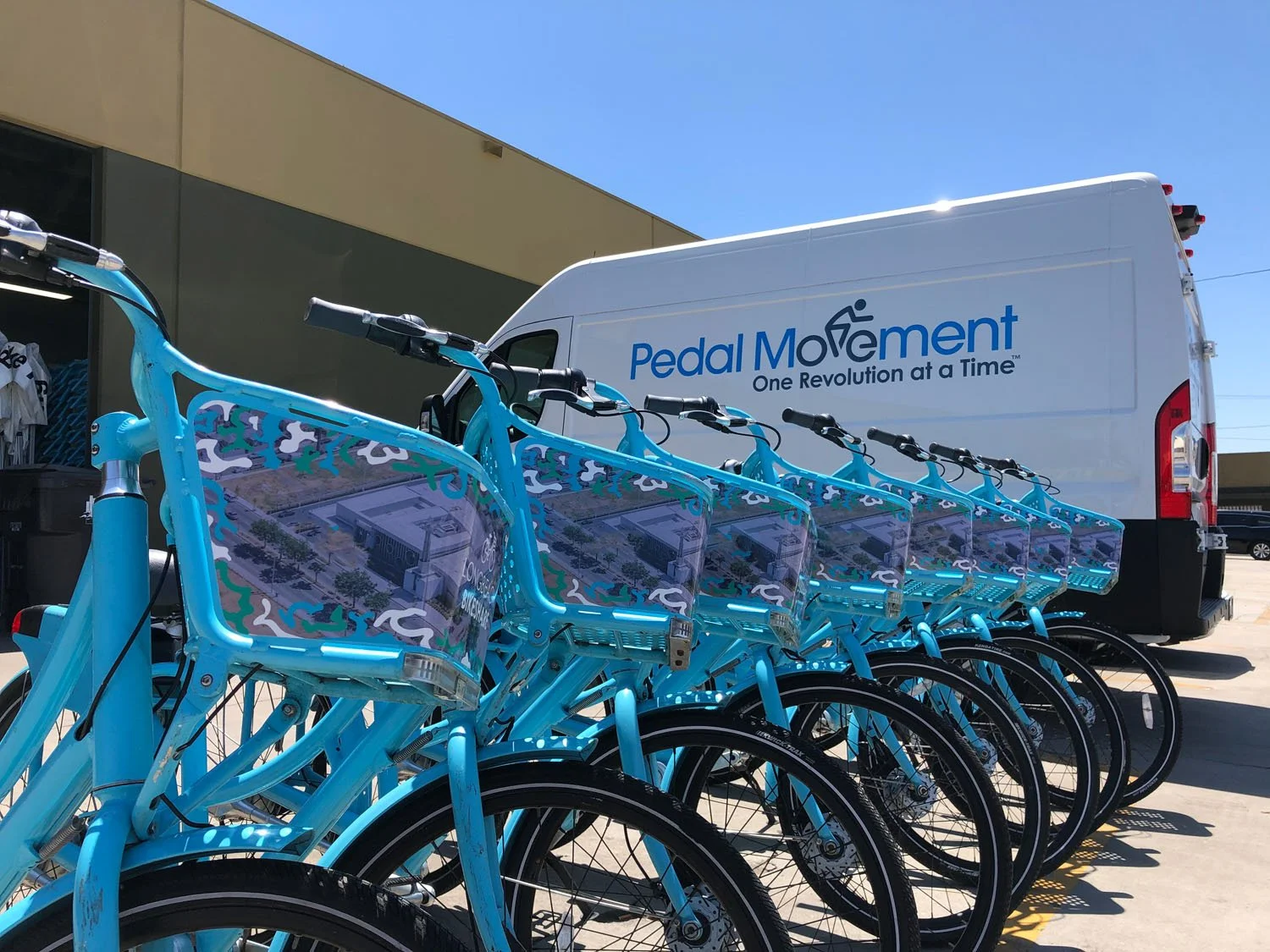 A row of blue bikes with baskets lined up in front of a white van with this words "Pedal Movement One Revolution at a Time" in an outdoor lot.