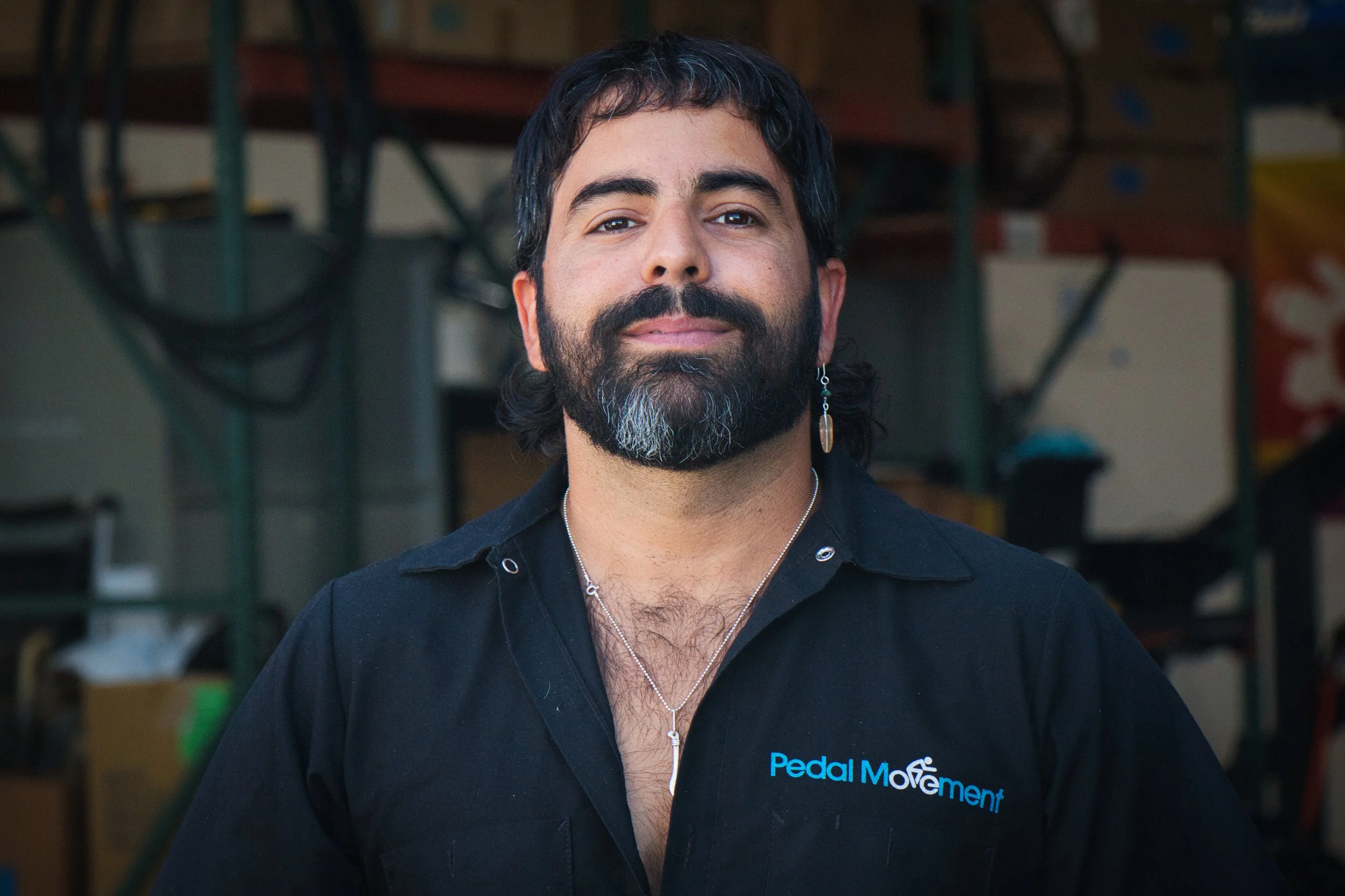 A man with dark hair, a beard with some gray, and a earring, wearing a black polo shirt with "Pedal Movement" embroidered on it, standing in front of a blurred background of garage or workshop tools.