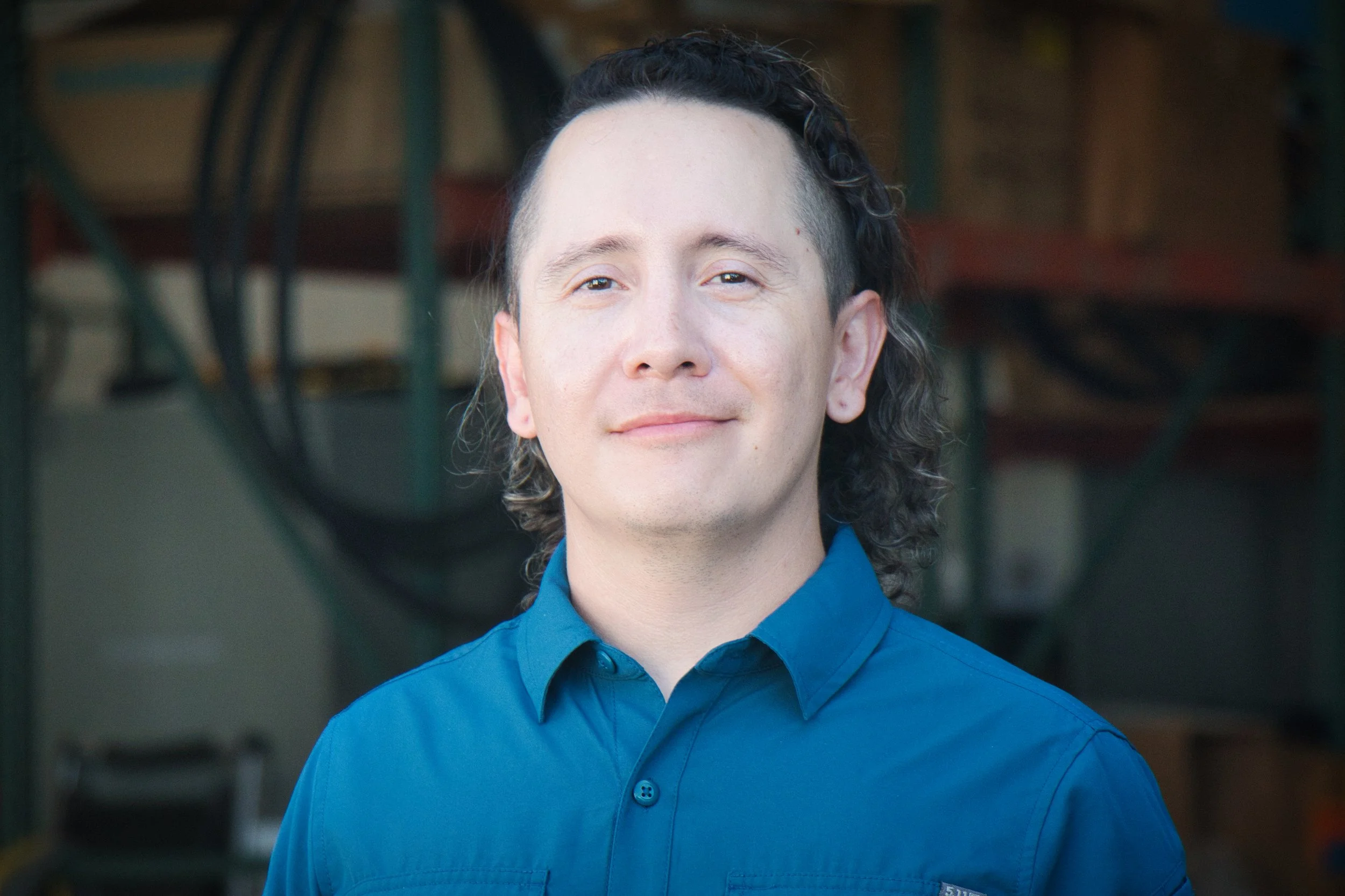 A man with black curly hair, wearing a blue button-up shirt, smiling softly, standing outdoors with blurred workshop in the background.