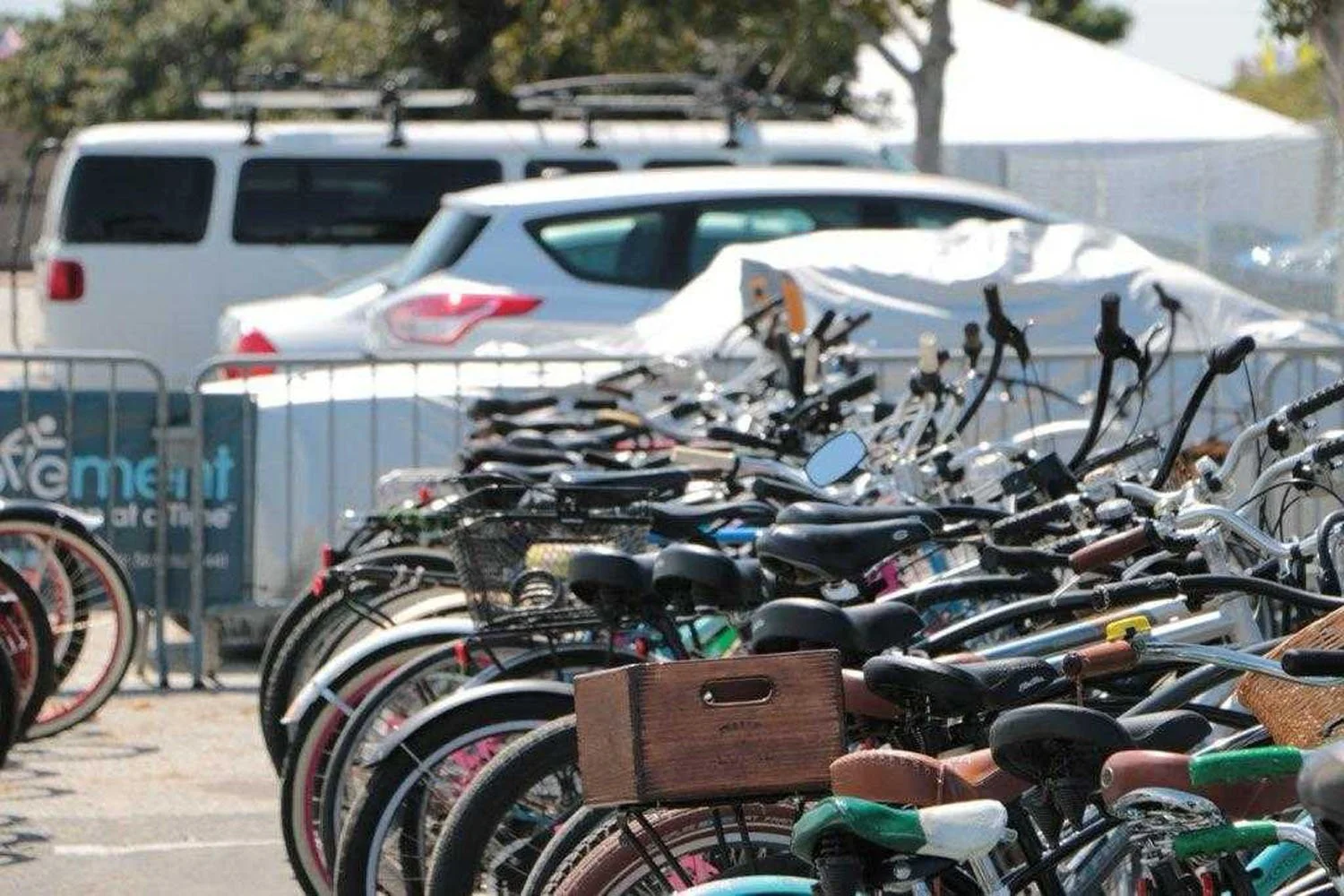 A bike parking lot with numerous bicycles, some with baskets, in the foreground and parked cars in the background.
