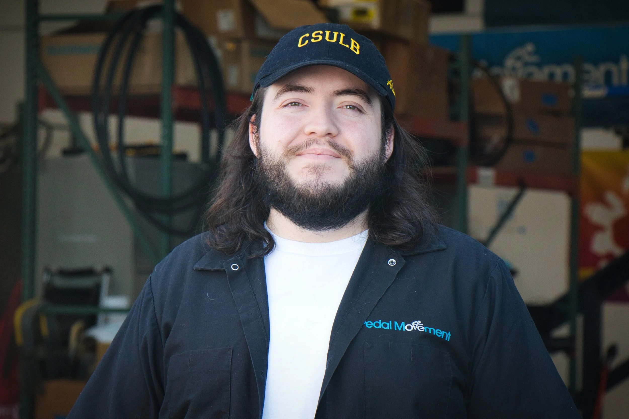 A man with long dark hair and a beard, smiling slightly, wearing a black jacket with the 'Pedal Movement' logo and a black cap with yellow lettering 'CSULB', standing in a workshop or storage room with shelves and equipment in the background.