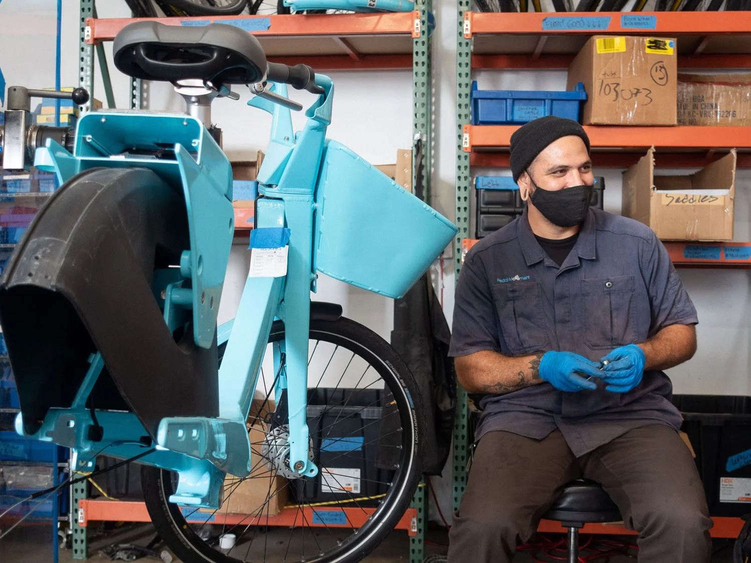 A person wearing a black mask and blue gloves sitting next to a partially assembled, bright blue bicycle inside a warehouse or workshop with shelving and boxes.