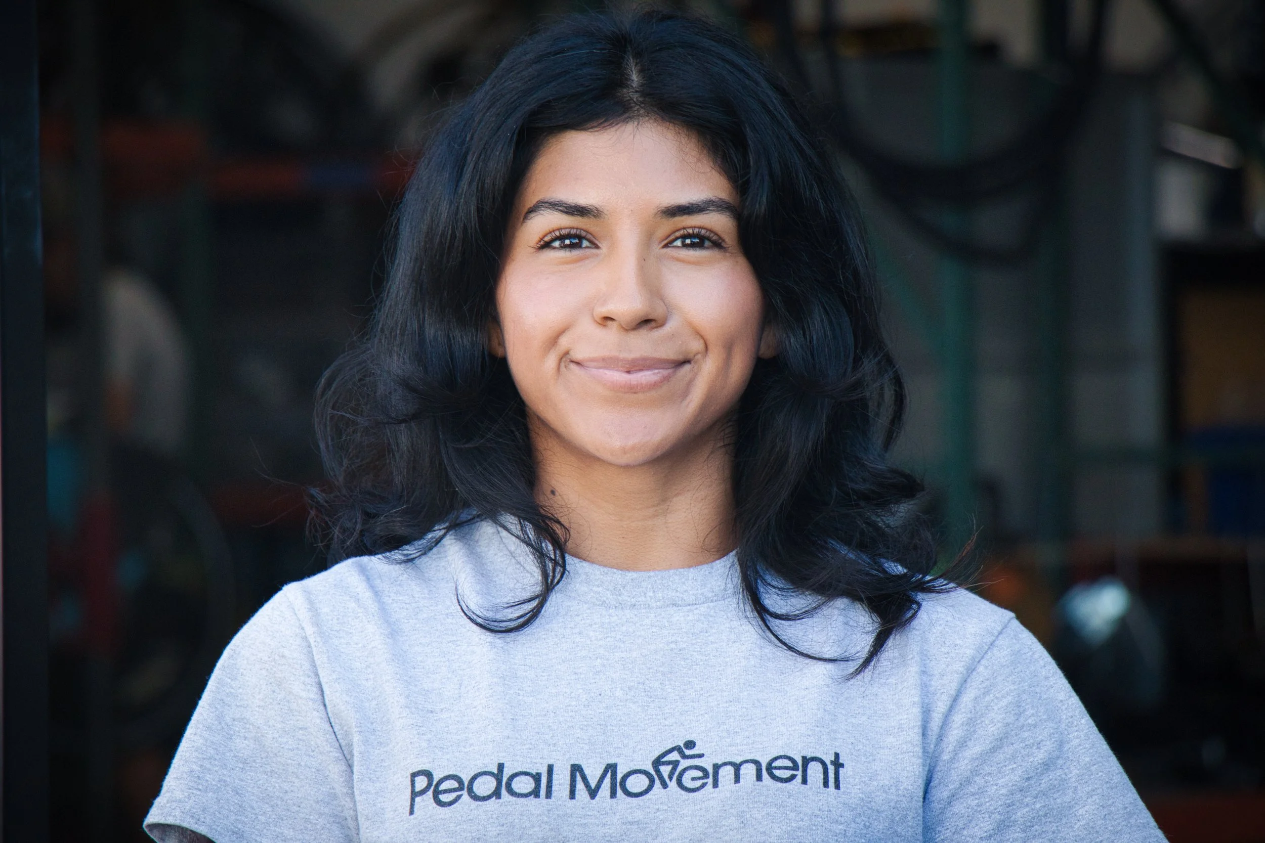 A woman with dark, wavy hair smiling at the camera, wearing a light gray t-shirt with the Pedal Movement logo.