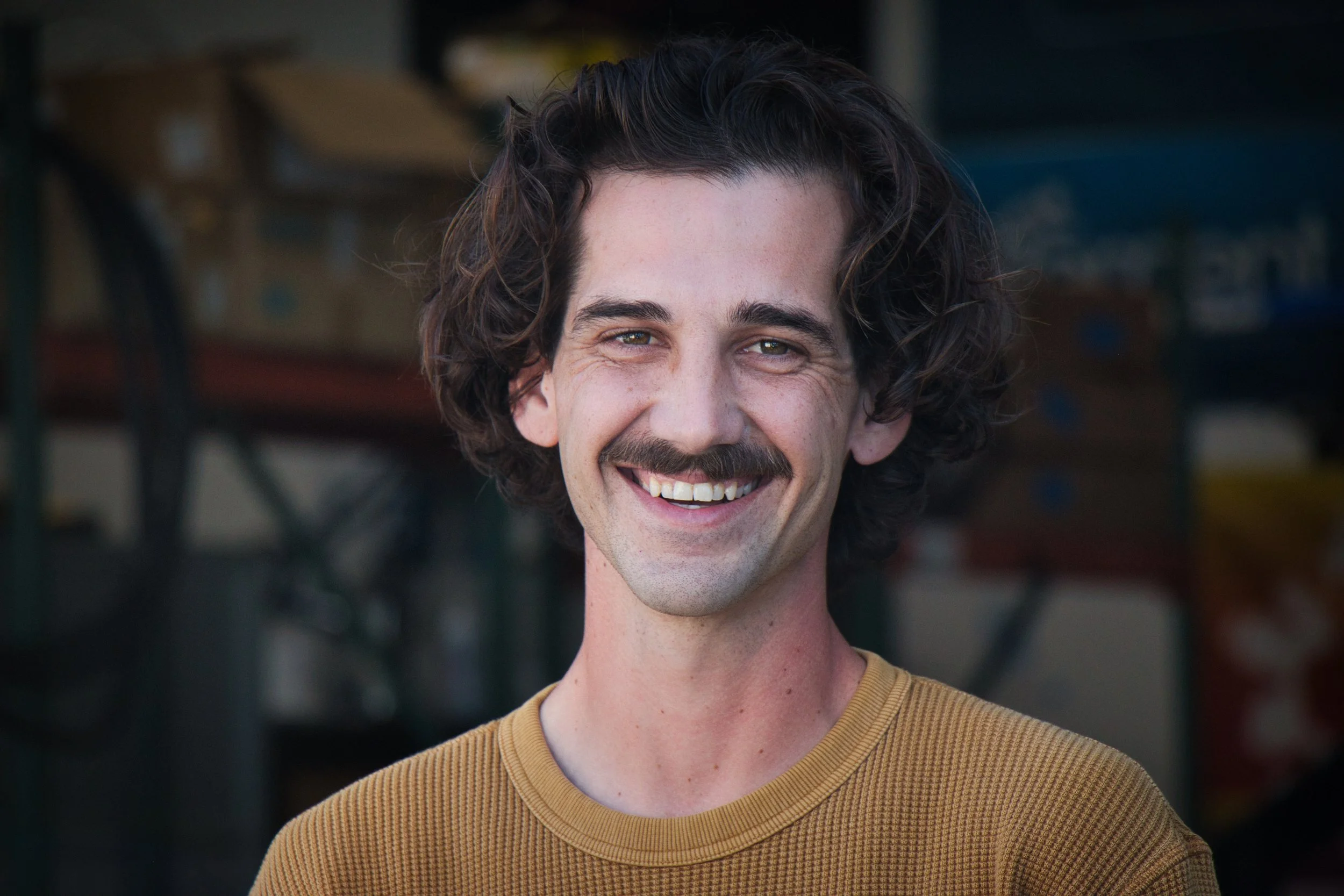 A young man with curly brown hair and a moustache, smiling in a close-up portrait. He's wearing a mustard yellow shirt, and the background is an industrial or workshop setting.