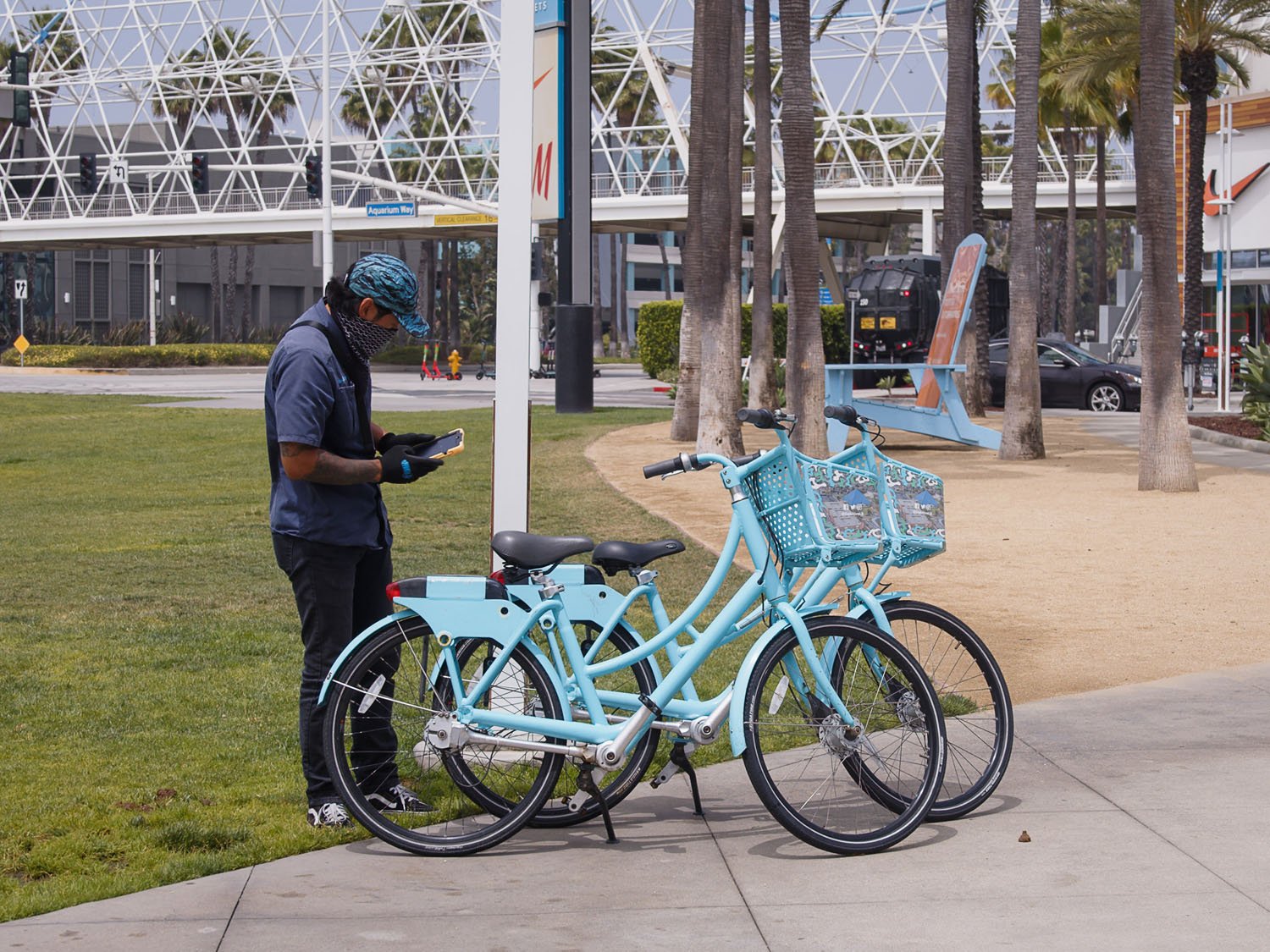 A person with a face covering standing next to a light blue bike with baskets in an outdoor park.