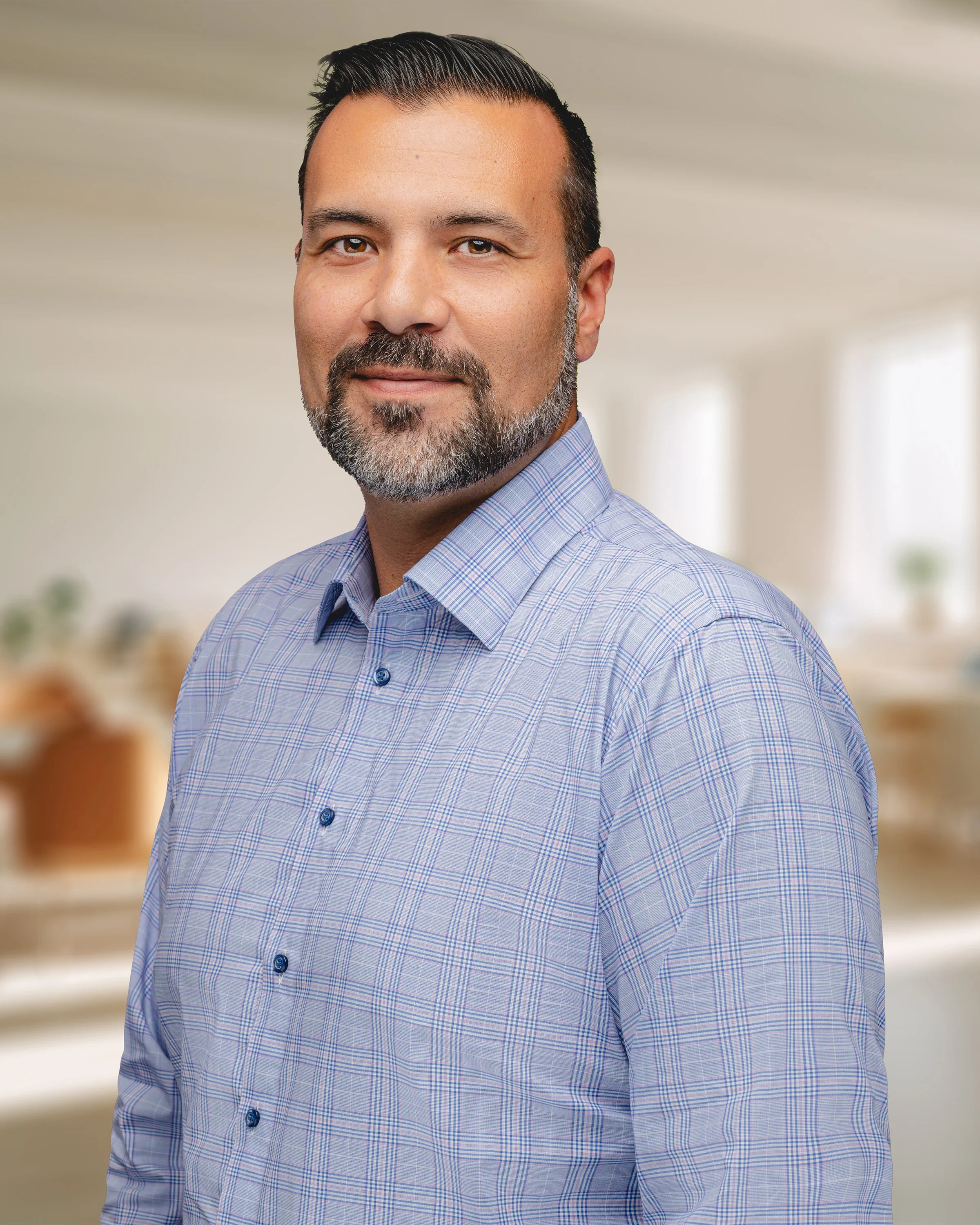 A man with dark hair and a beard wearing a light blue checkered dress shirt, standing in a bright indoor space that appears to be an office or a home.