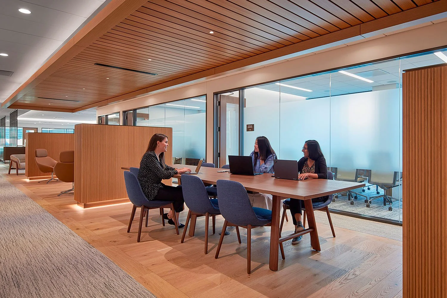Three women working on laptops in a modern, well-lit conference room with wooden floors and glass walls.
