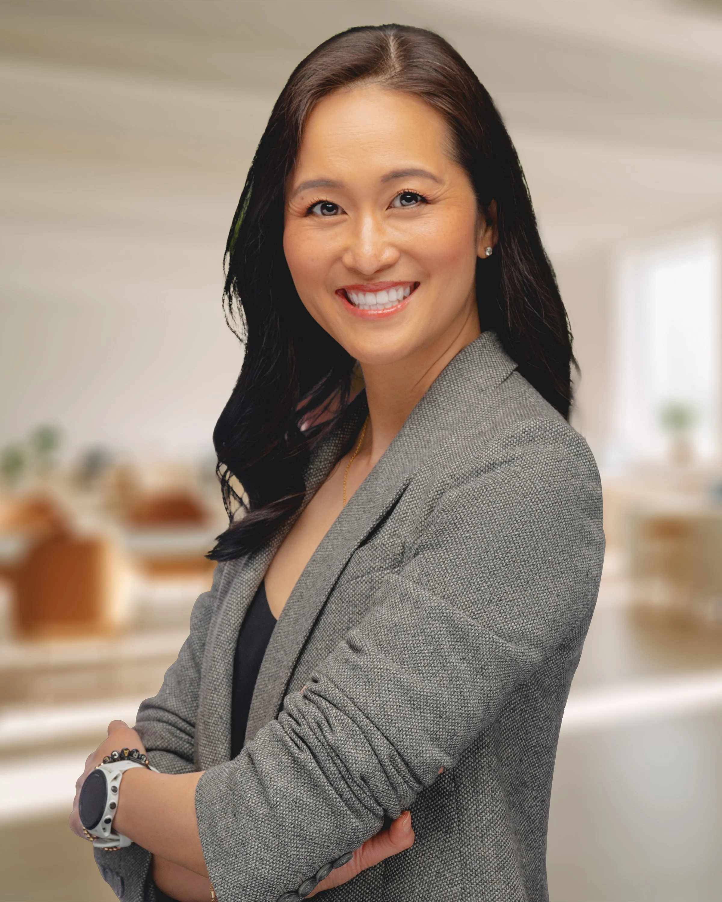 A woman with black hair wearing a gray blazer smiling with arms crossed in an office setting.