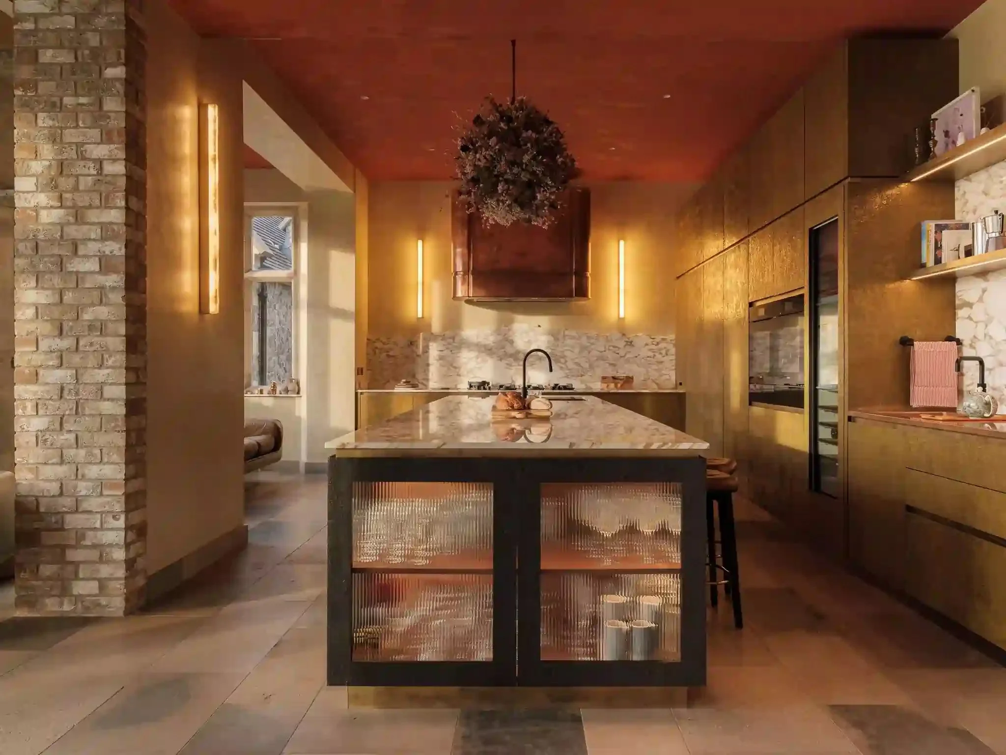 Modern kitchen with gold cabinets, marble countertops, a kitchen island with textured glass cabinets, exposed brick wall, and warm lighting.