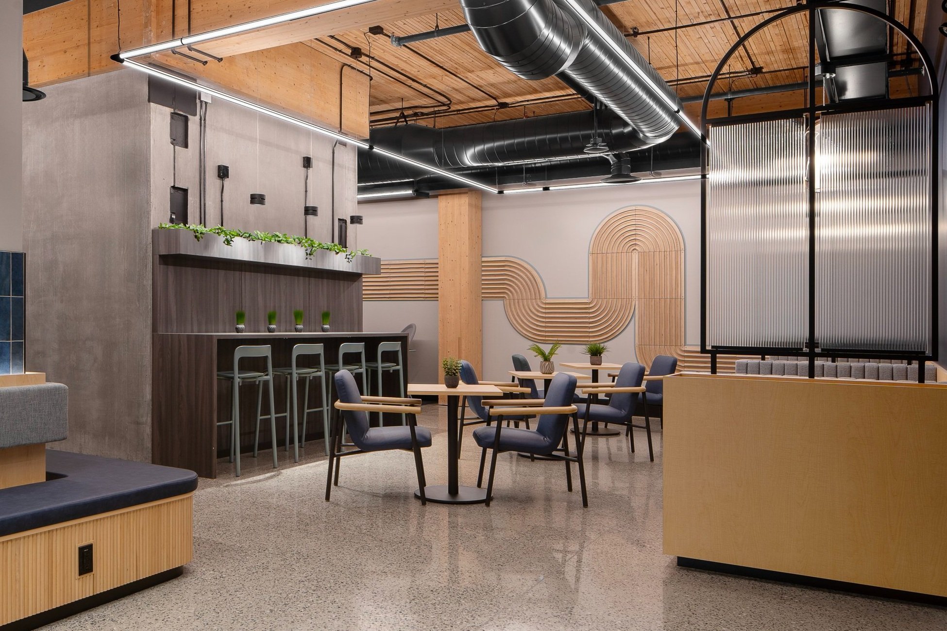 Modern office lounge area with tables, chairs, plants, decorative wall art, and industrial ceiling design.