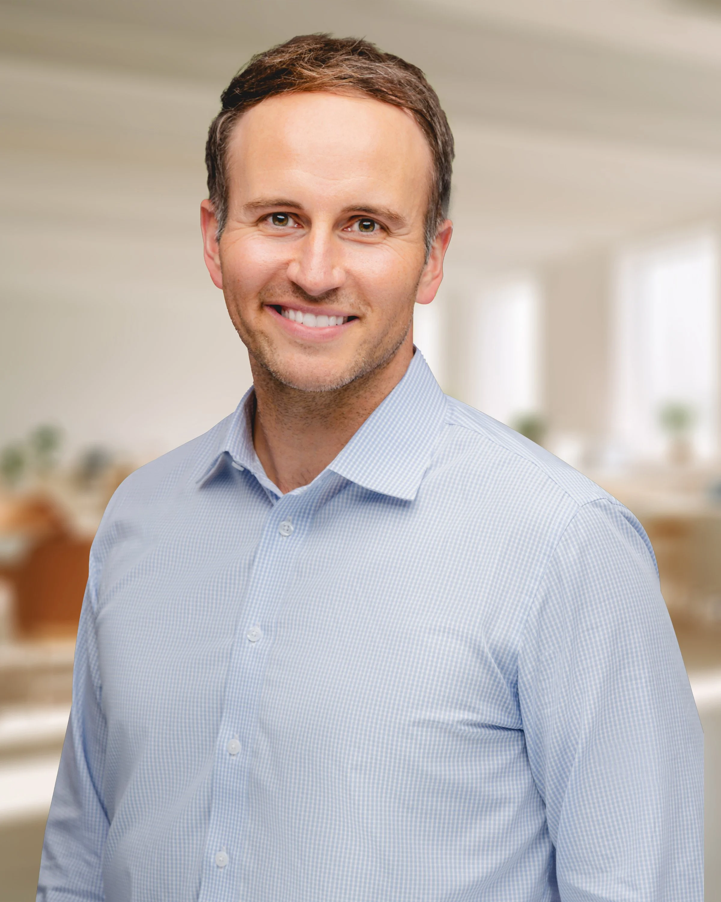 A man with brown hair, smiling, wearing a light blue collared shirt, standing in a bright, modern kitchen.