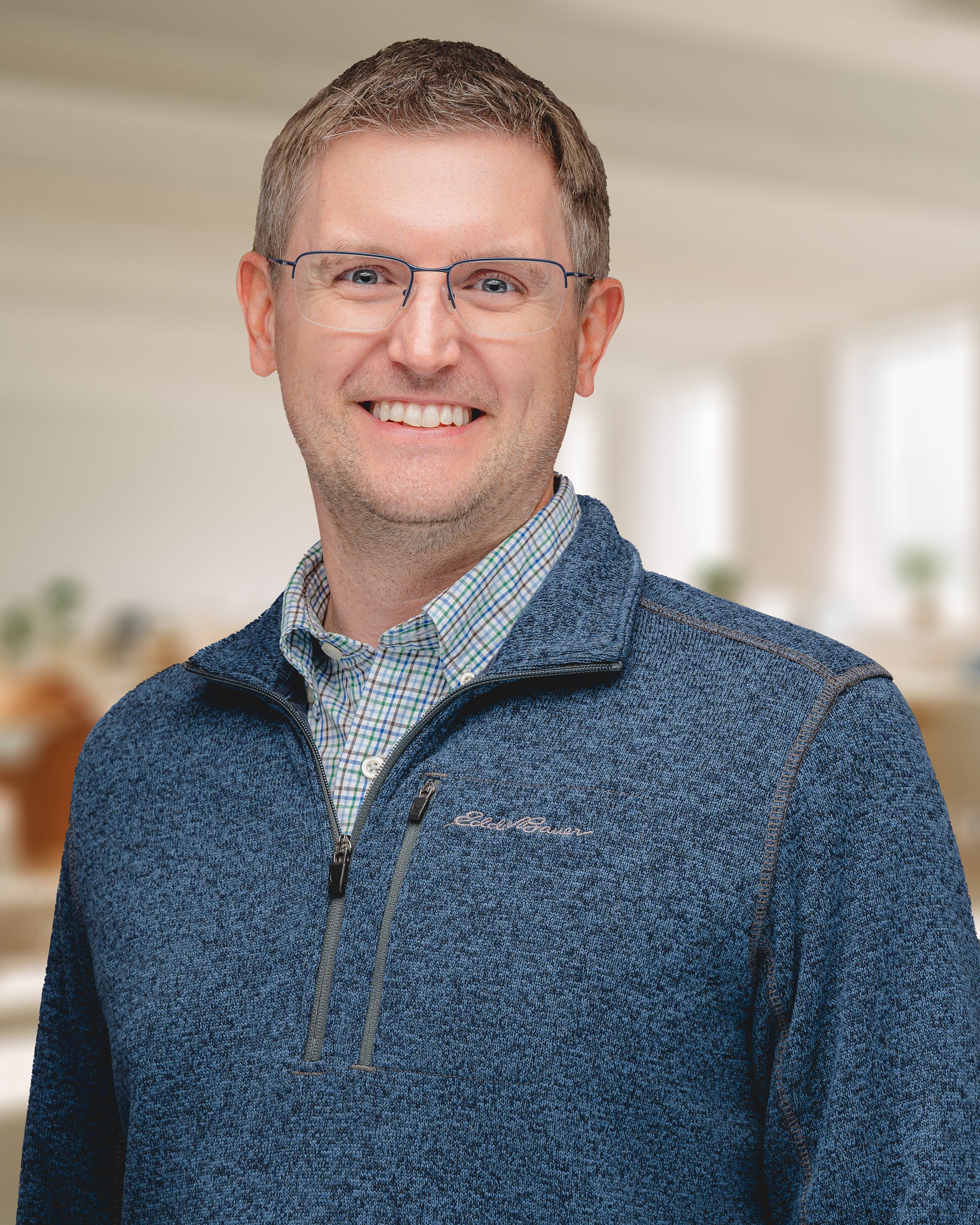 A smiling man with glasses, short brown hair, wearing a blue fleece jacket over a plaid shirt, in a bright indoor setting.