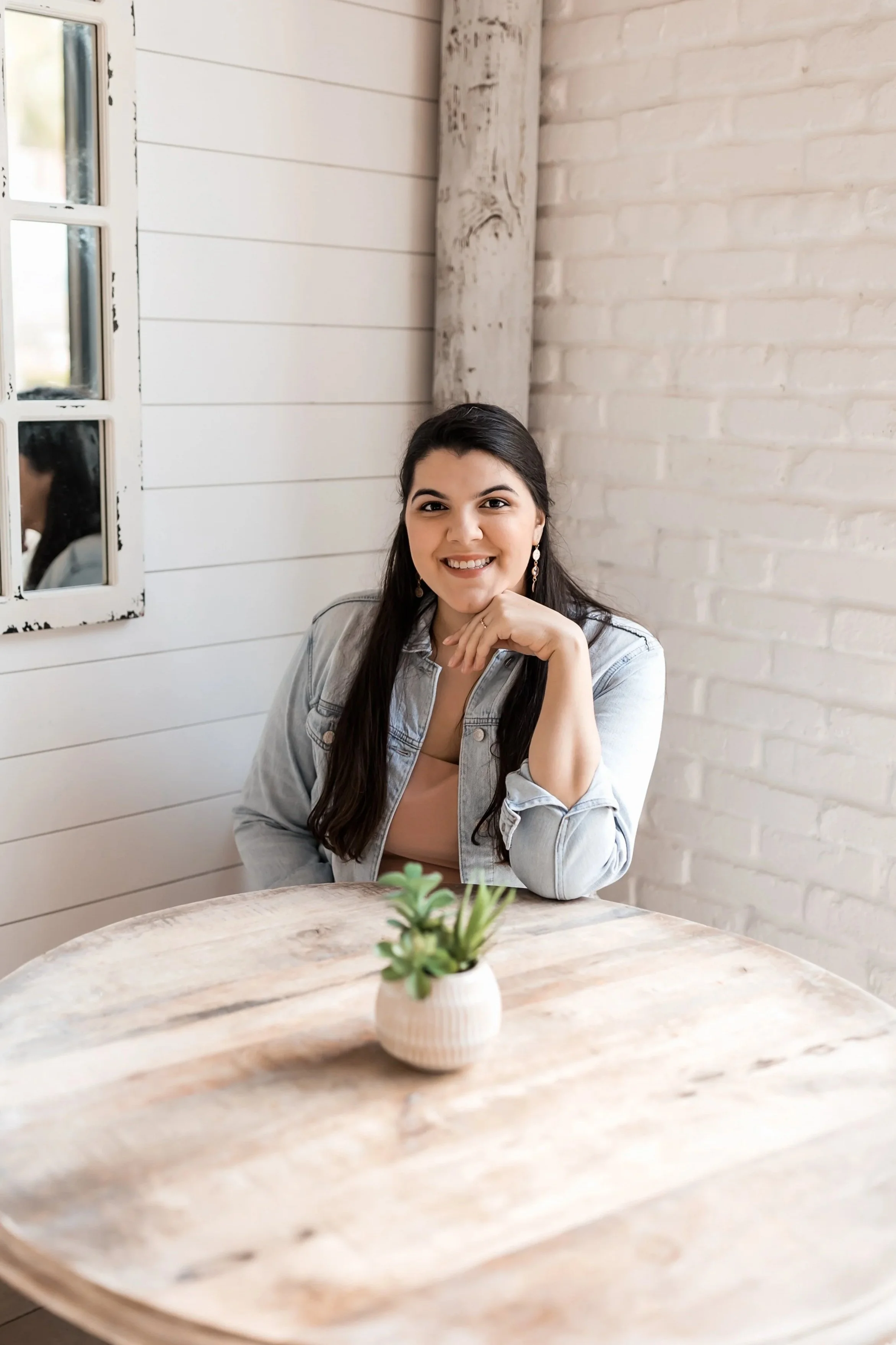 A woman with long dark hair, wearing earrings, a light pink top, and a denim jacket, smiling and resting her chin on her right hand, sitting at a light wooden table with a small potted succulent plant on it, inside a cozy room with white shiplap walls and a mirror on the wall.