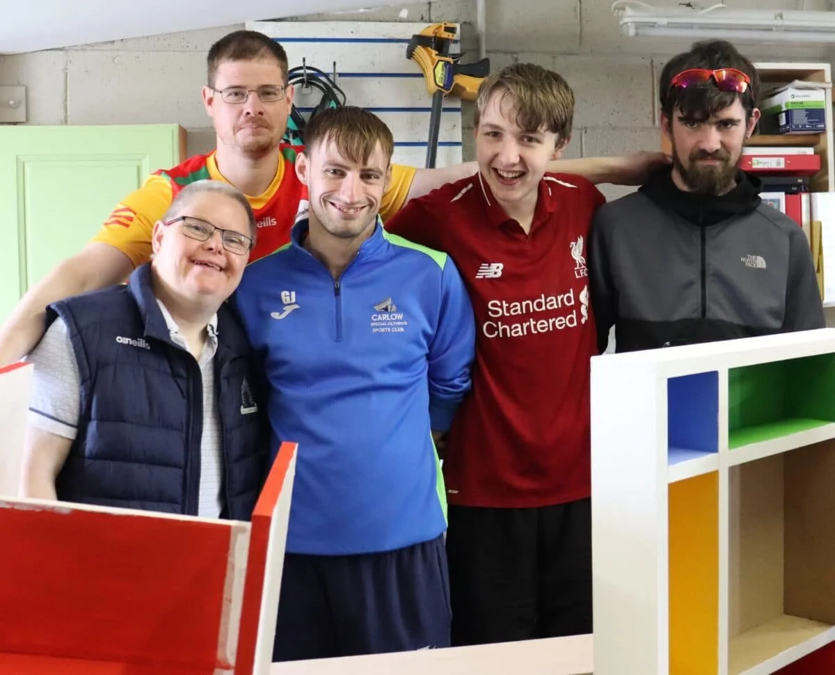 Five men posing together indoors, smiling and standing close, with tools and shelves in the background. One man is wearing a Liverpool FC jersey, and two others are wearing blue jackets with sports club logos.