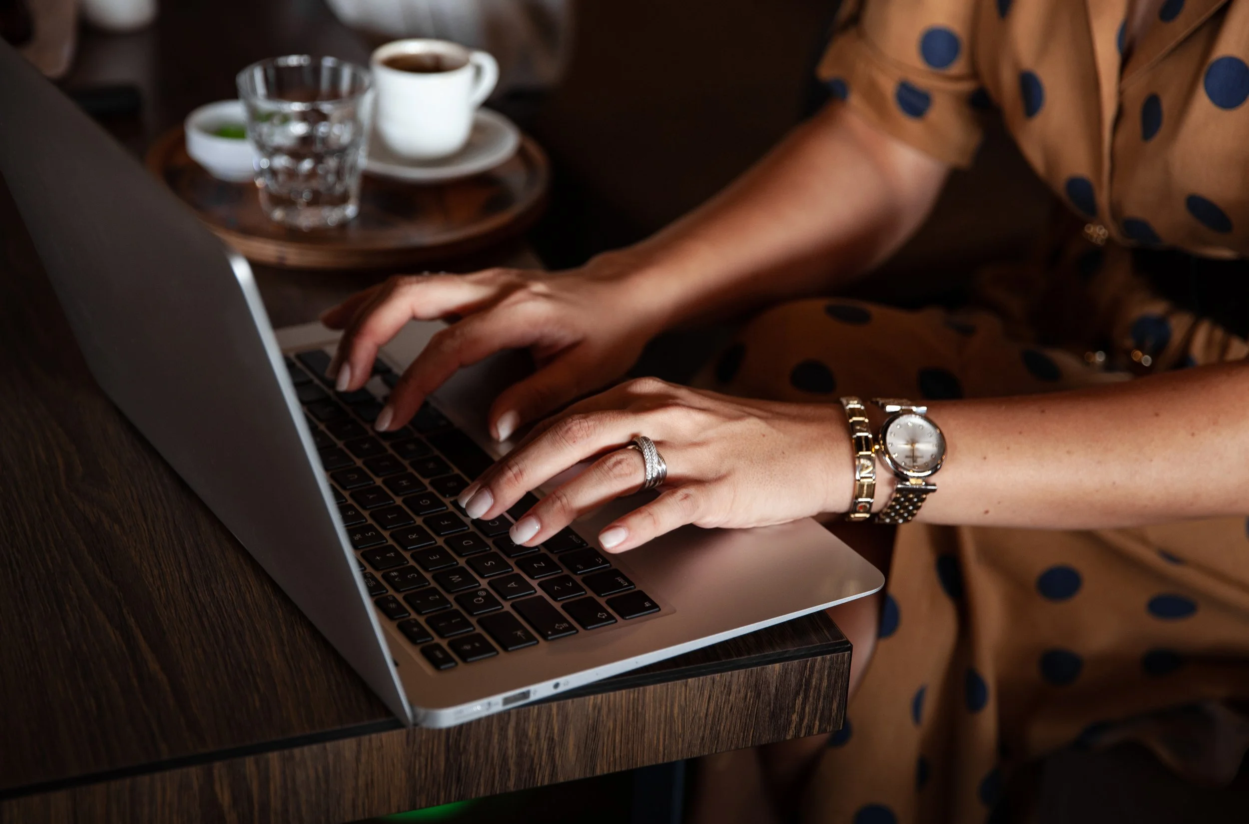 Person typing on a laptop with a watch and rings on their fingers, sitting at a wooden table with a cup of coffee and glass of water in the background.