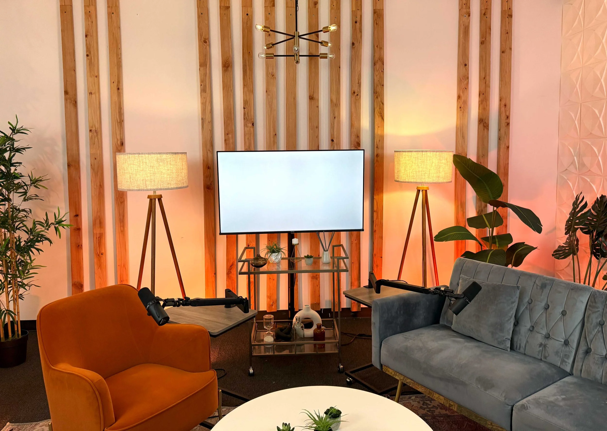 Modern living room with a wooden wall, two tripod lamps, a large TV, two podcasts microphones, a gray couch, an orange armchair, plants, and a small white table.