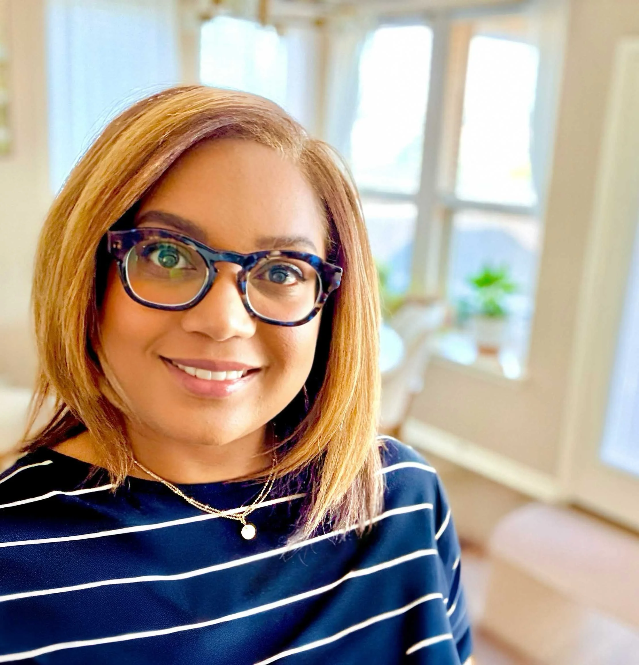 This is Francine Henry - A pretty black woman with shoulder-length auburn hair and glasses, smiling inside a bright room with large windows and potted plants in the background.