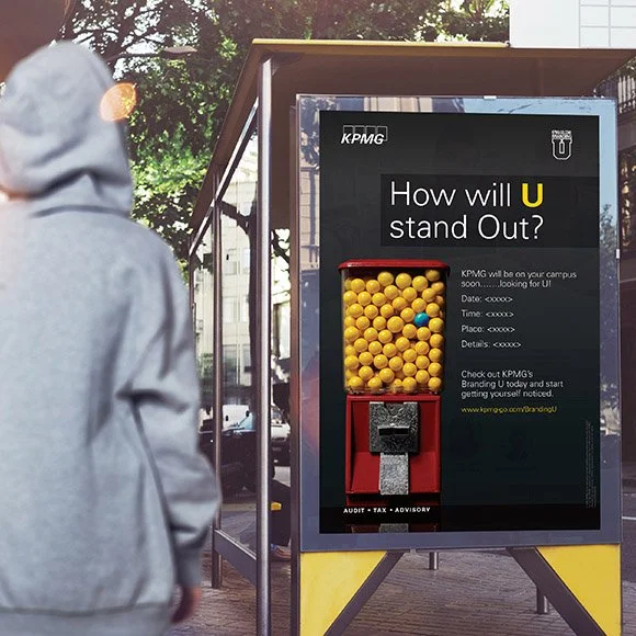 A person wearing a gray hoodie walking past a bus stop with an advertisement for KPMG featuring a yellow gumball machine and the text "How will you stand out?" on a bus shelter poster.