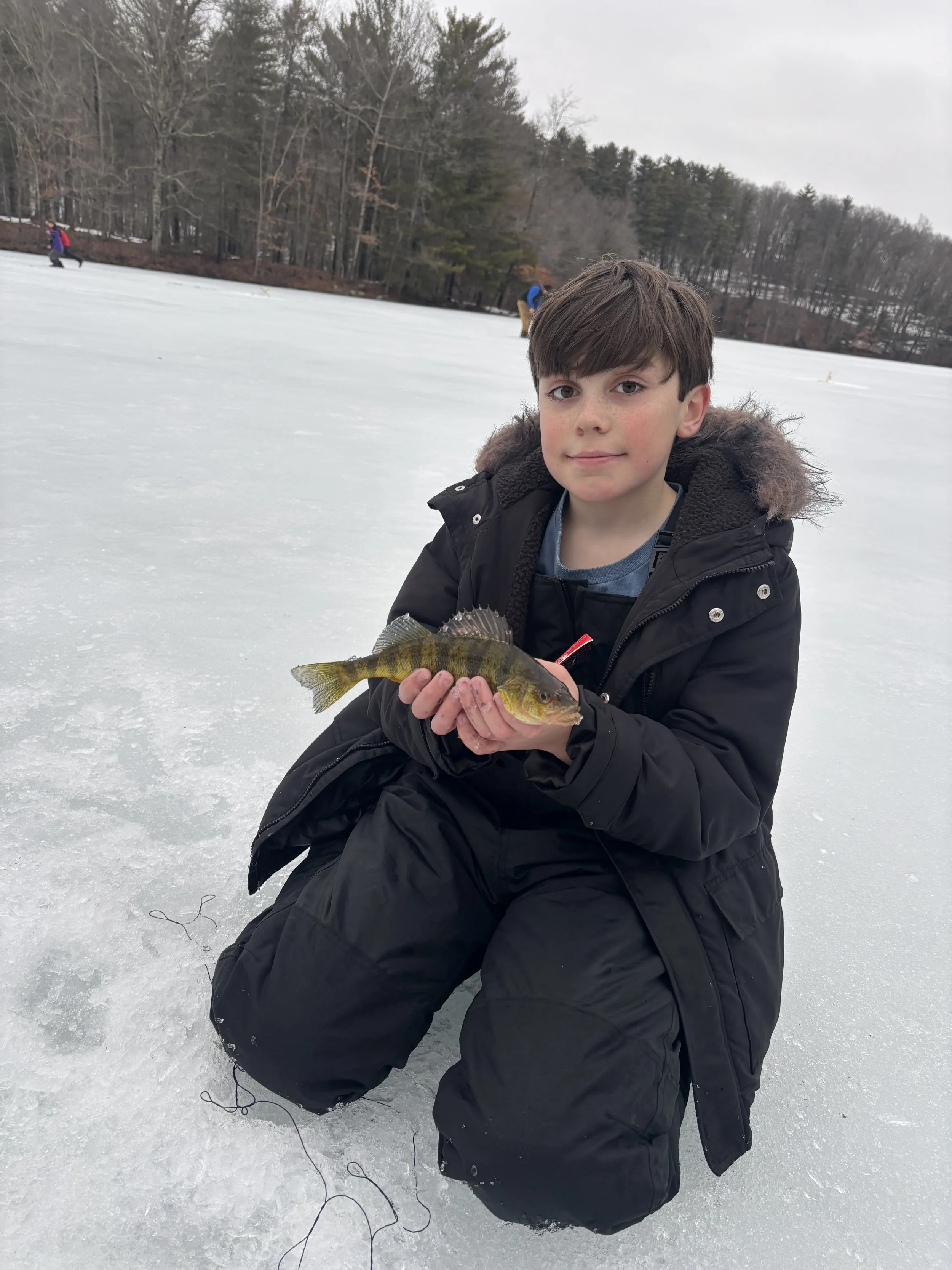 A boy kneeling on ice while holding a fish he caught, with a frozen lake and trees in the background.