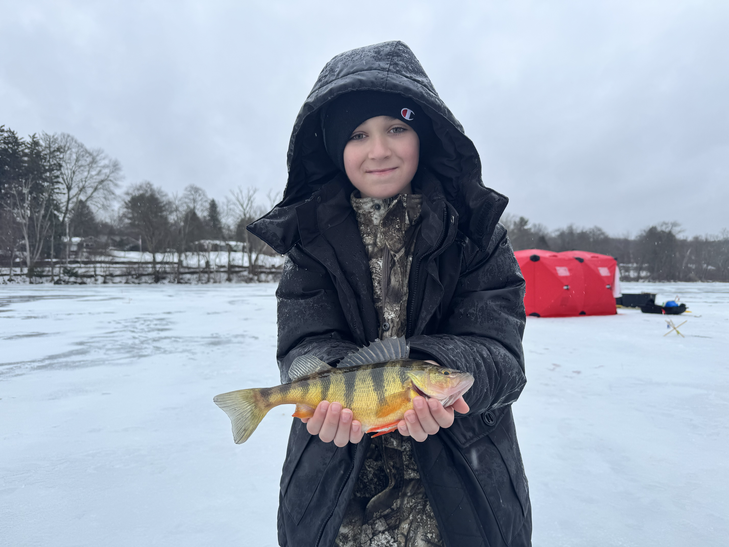 A young boy dressed in winter clothing, holding a fish on a snow-covered ice lake during winter.