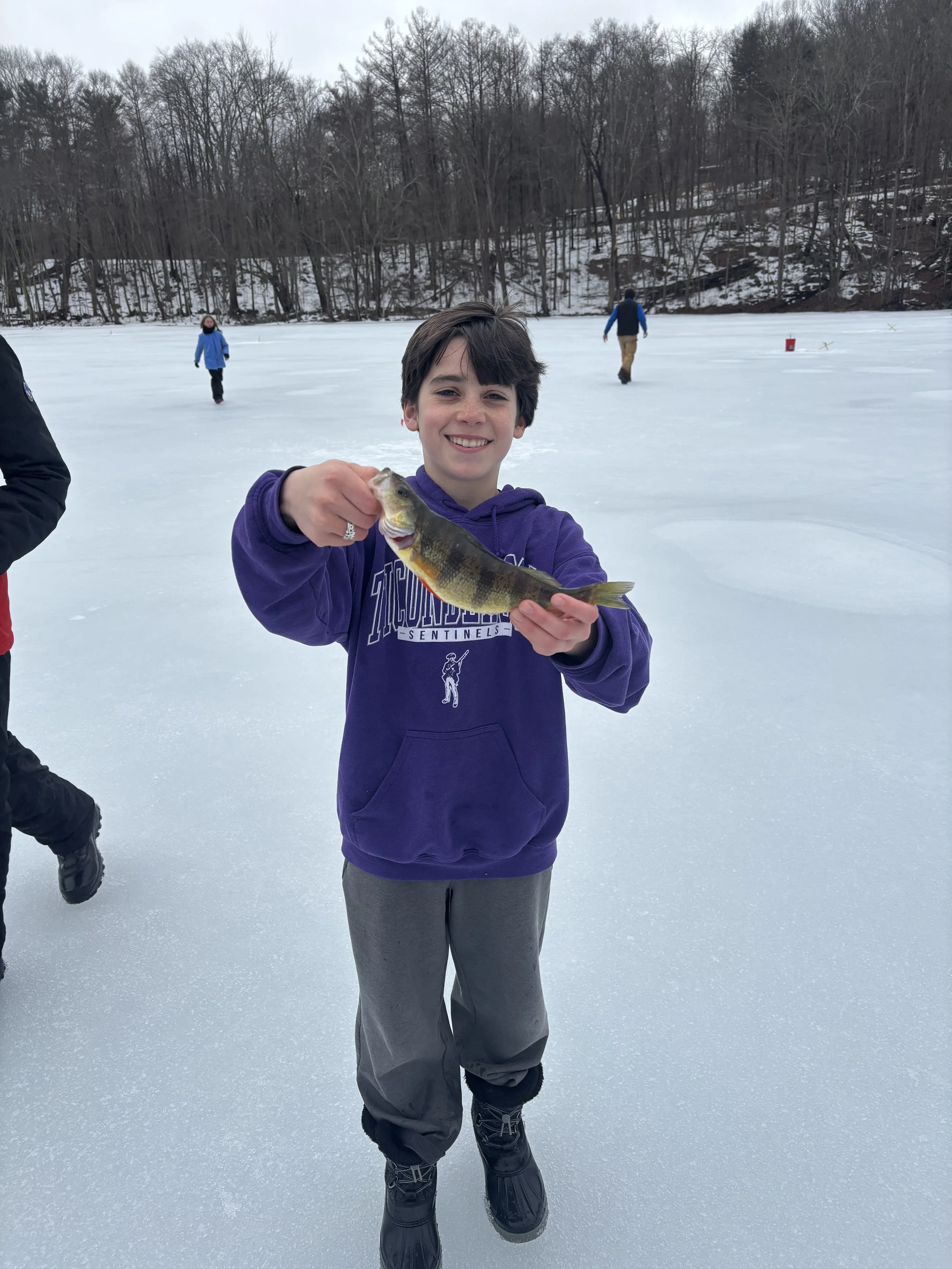 A boy is on a frozen lake holding a fish he caught, smiling at the camera. He is dressed in a purple hoodie, gray pants, and black winter boots. Other people can be seen ice fishing in the background, with leafless trees and snow-covered ground behin