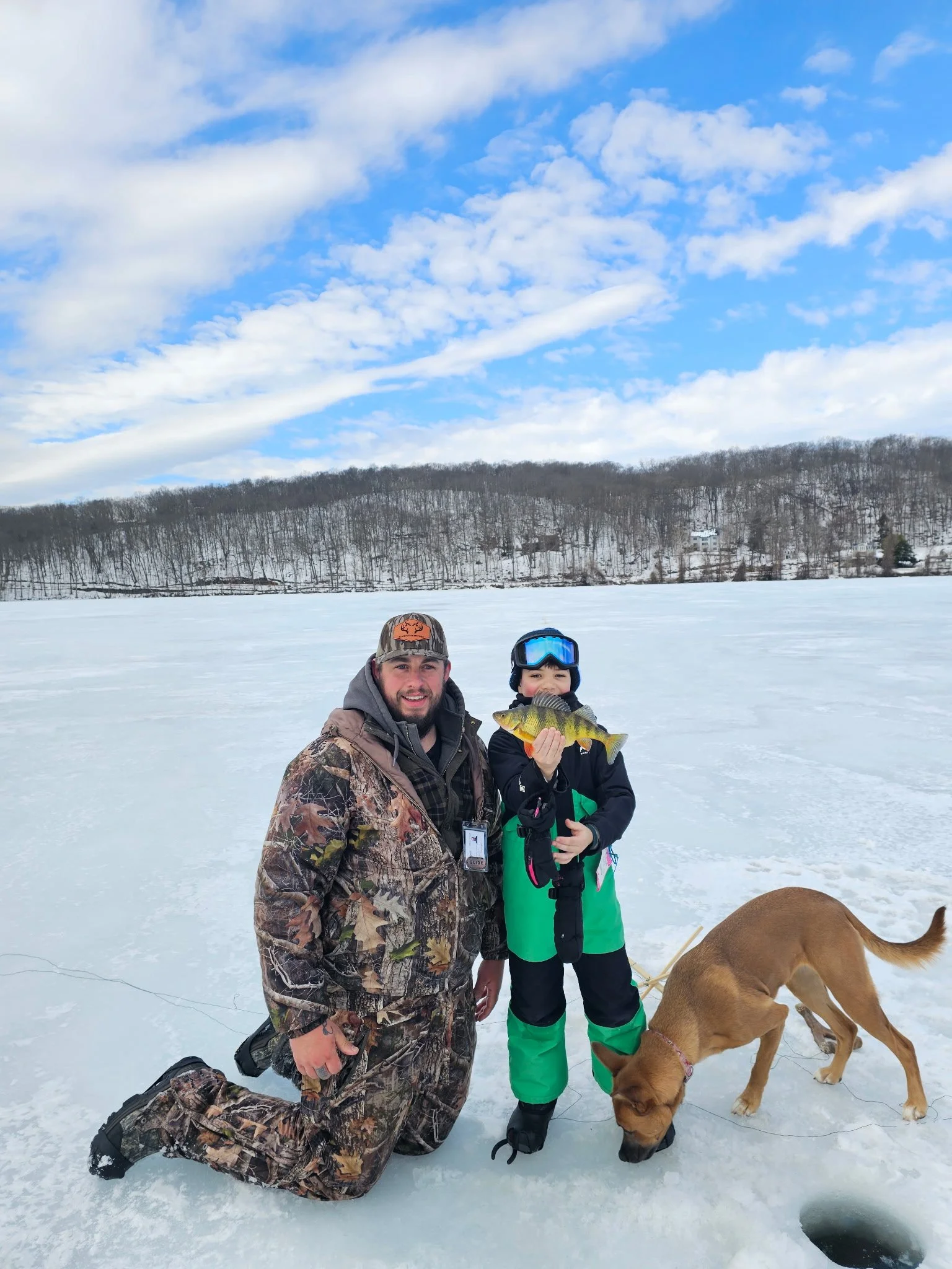 A man and a boy on an icy lake, with the boy holding a fish, a dog sniffing the ice, and snow-covered trees in the background.