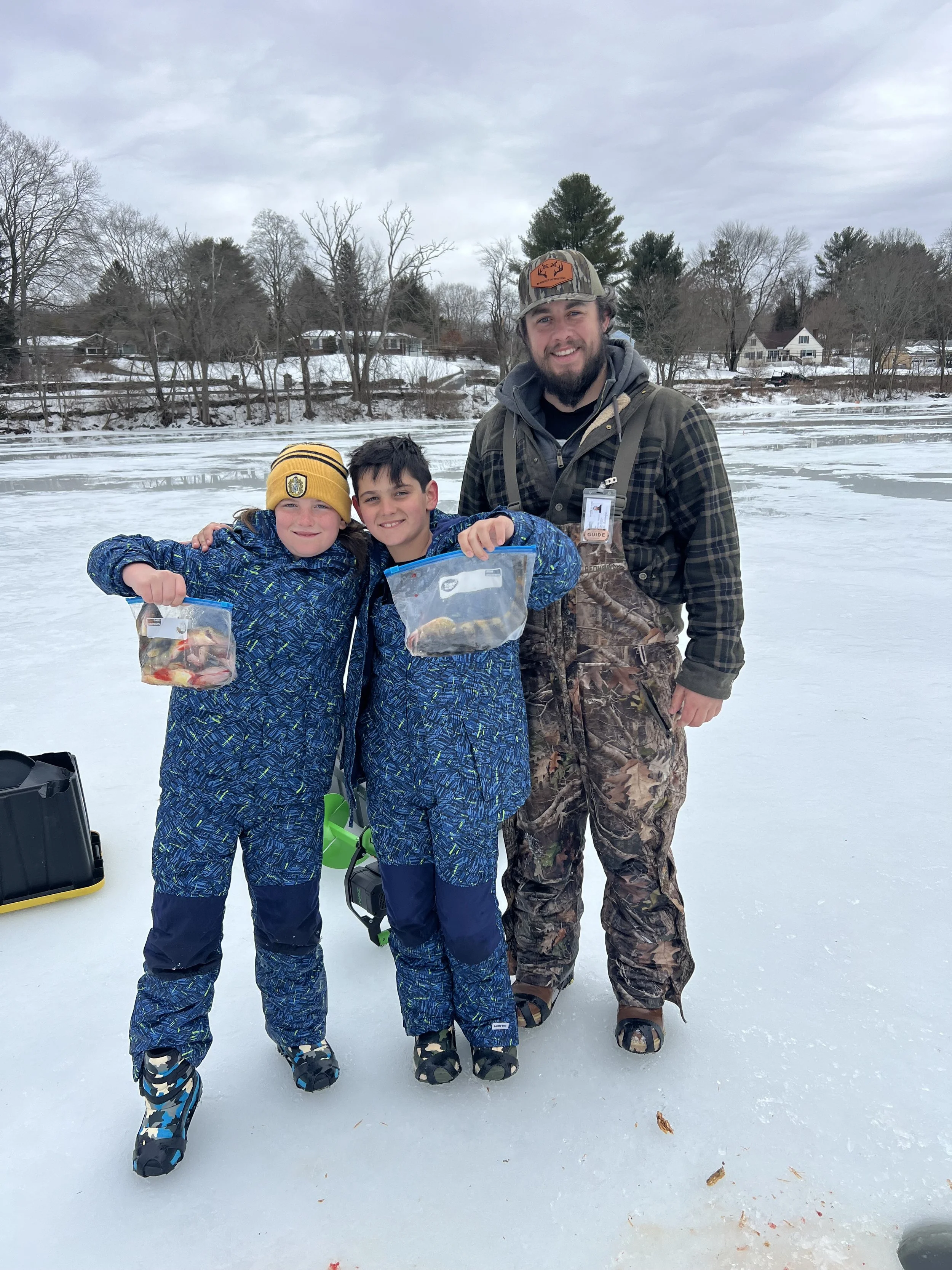 Three people, two children and one adult, standing on a frozen lake holding plastic bags with fish, dressed in winter clothing, with a snowy landscape and trees in the background.