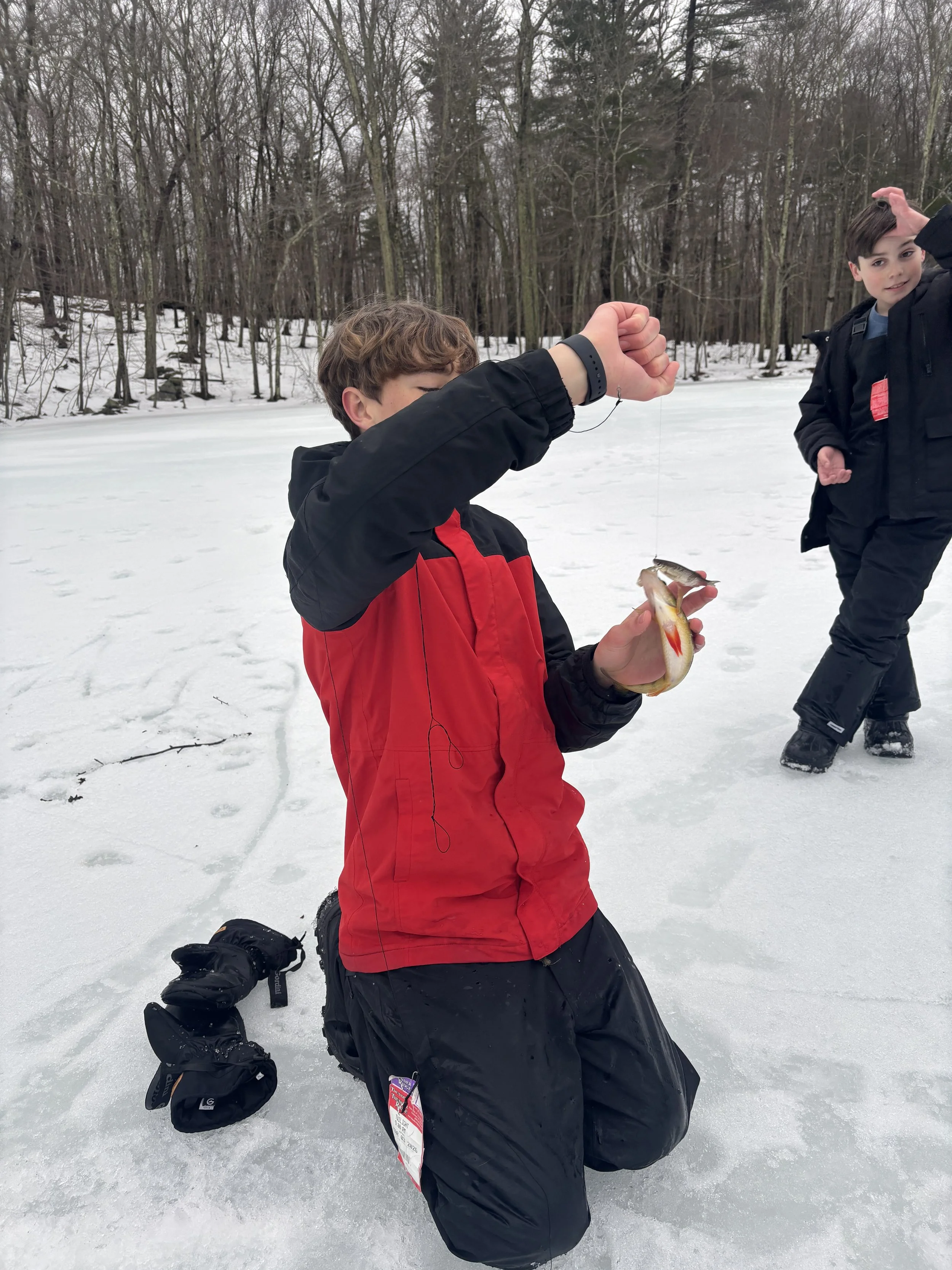 A boy dressed in a red and black winter coat kneeling on snow while holding a fishing line with a fish hanging from it, with another boy standing in the background in a snowy outdoor setting with trees.
