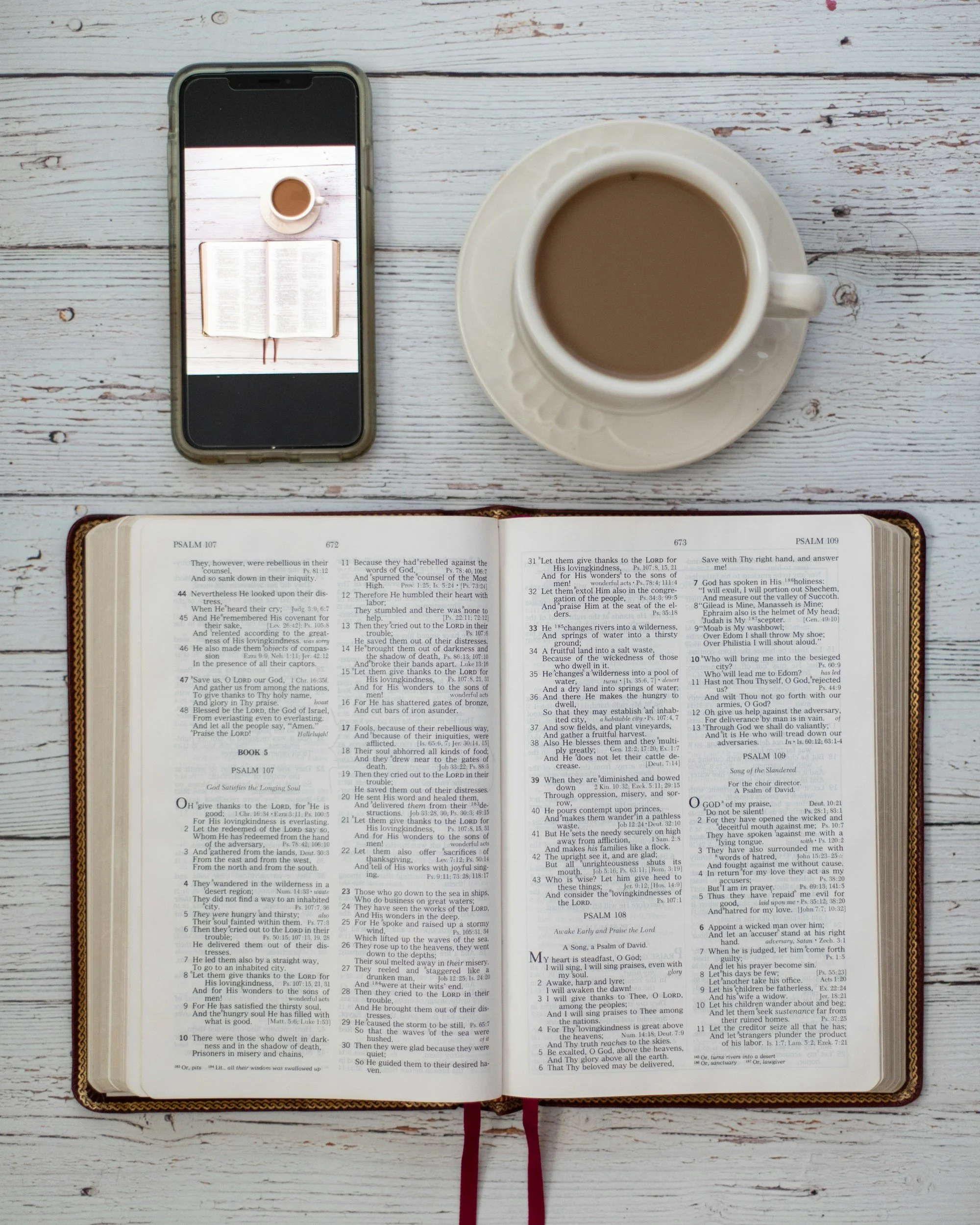 Open Bible with Psalm passages, a smartphone displaying a top-down view of a mug of coffee, and a saucer on a white wooden table.