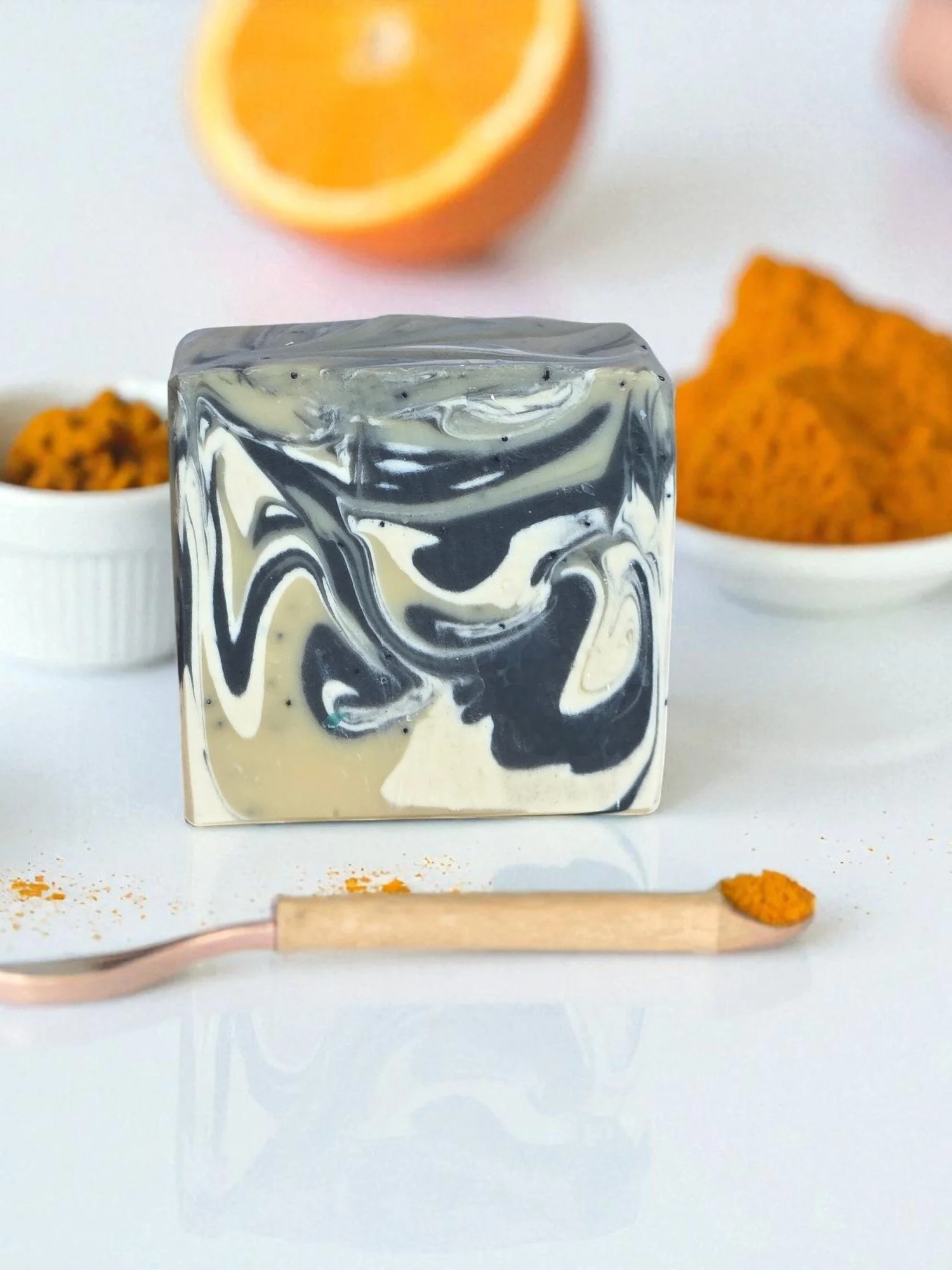 A bar of soap with black and white marbled swirl design, surrounded by bowls of tea and spices, with a scoop of turmeric in the foreground.