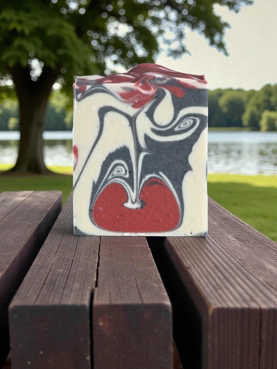A square bar of marble-patterned soap with red, black, and white swirls, placed on a white surface with a knife and peeled apple slices nearby, and a bowl of red apples in the background.