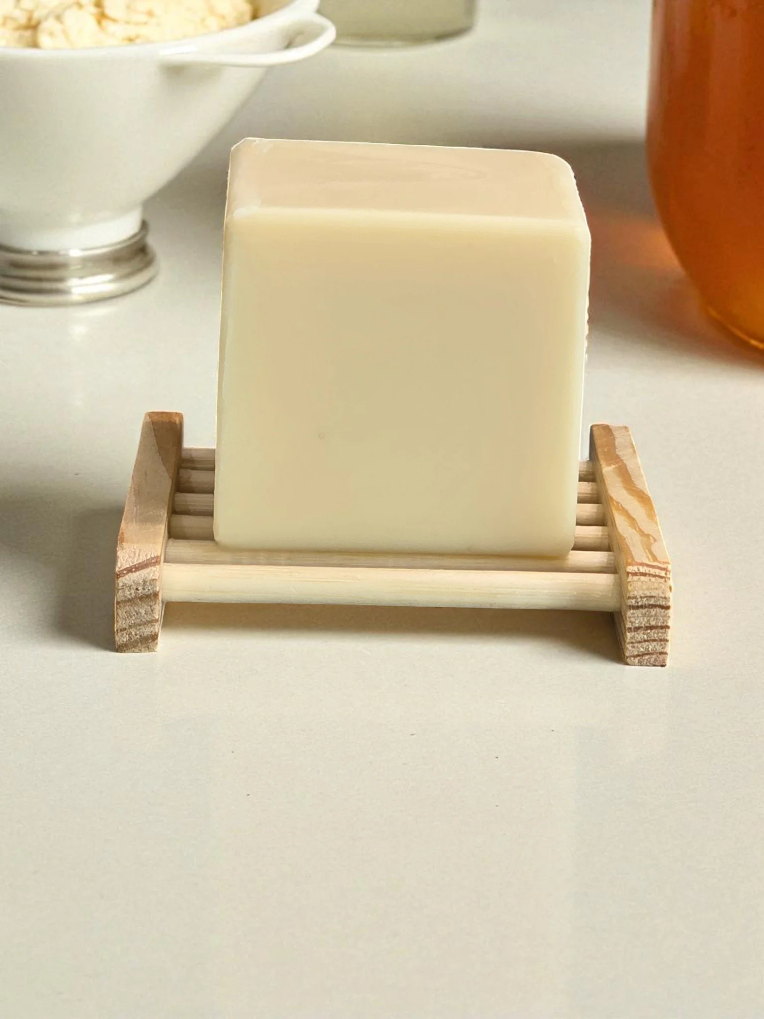 A lotion bar on a wooden soap dish with a vase of pink flowers in the background.