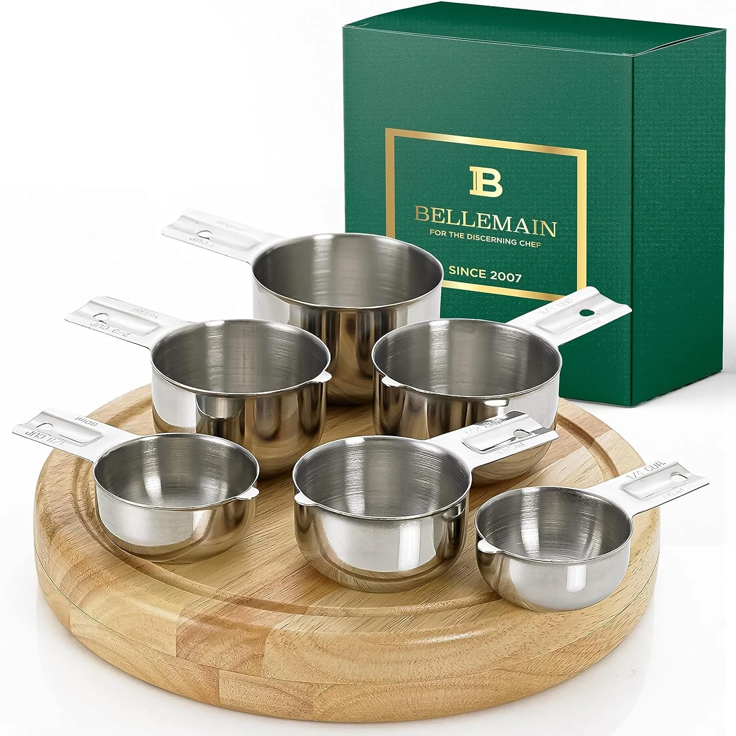 Set of six stainless steel measuring cups on a round wooden tray with a green box in the background labeled Belmain, for measuring ingredients.