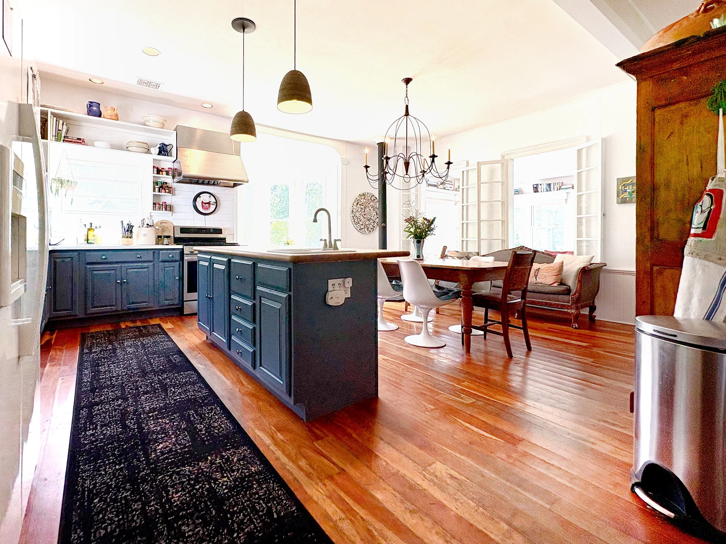 Open-concept kitchen and dining area with white walls, large windows, blue kitchen cabinets, wooden dining table, hanging light fixtures, and hardwood floors.