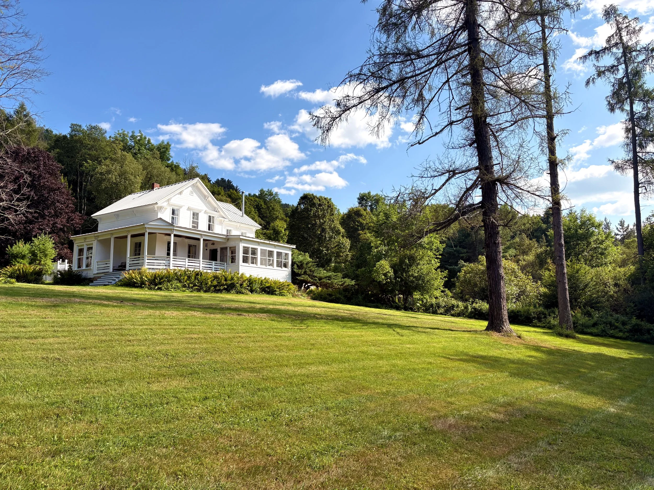 A white house with a porch sits on a grassy hill surrounded by trees under a blue sky with scattered clouds.