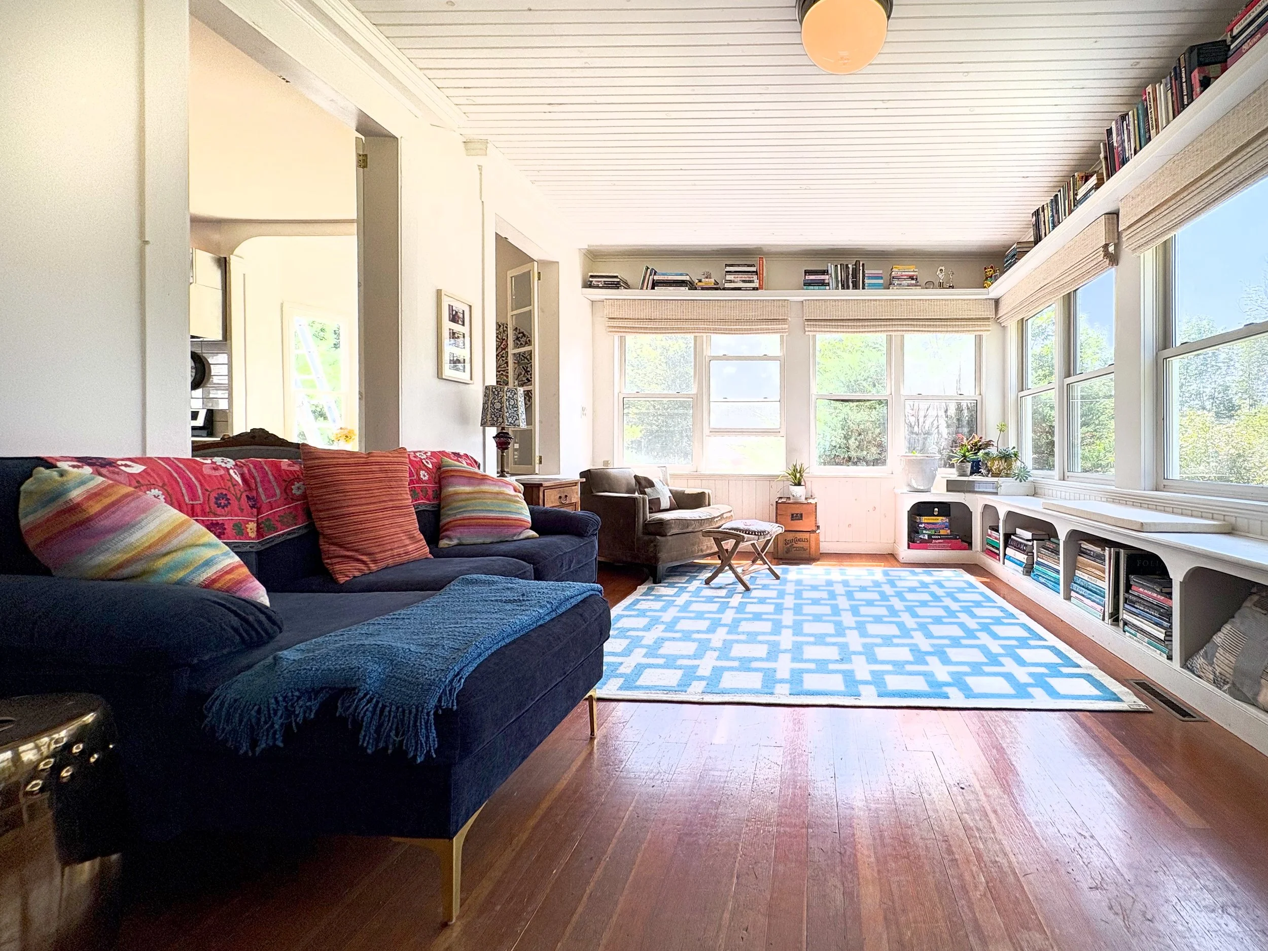 Bright living room with large windows, a blue couch with colorful pillows, a patterned blue and white rug, bookshelves along the wall, and a small wooden table.