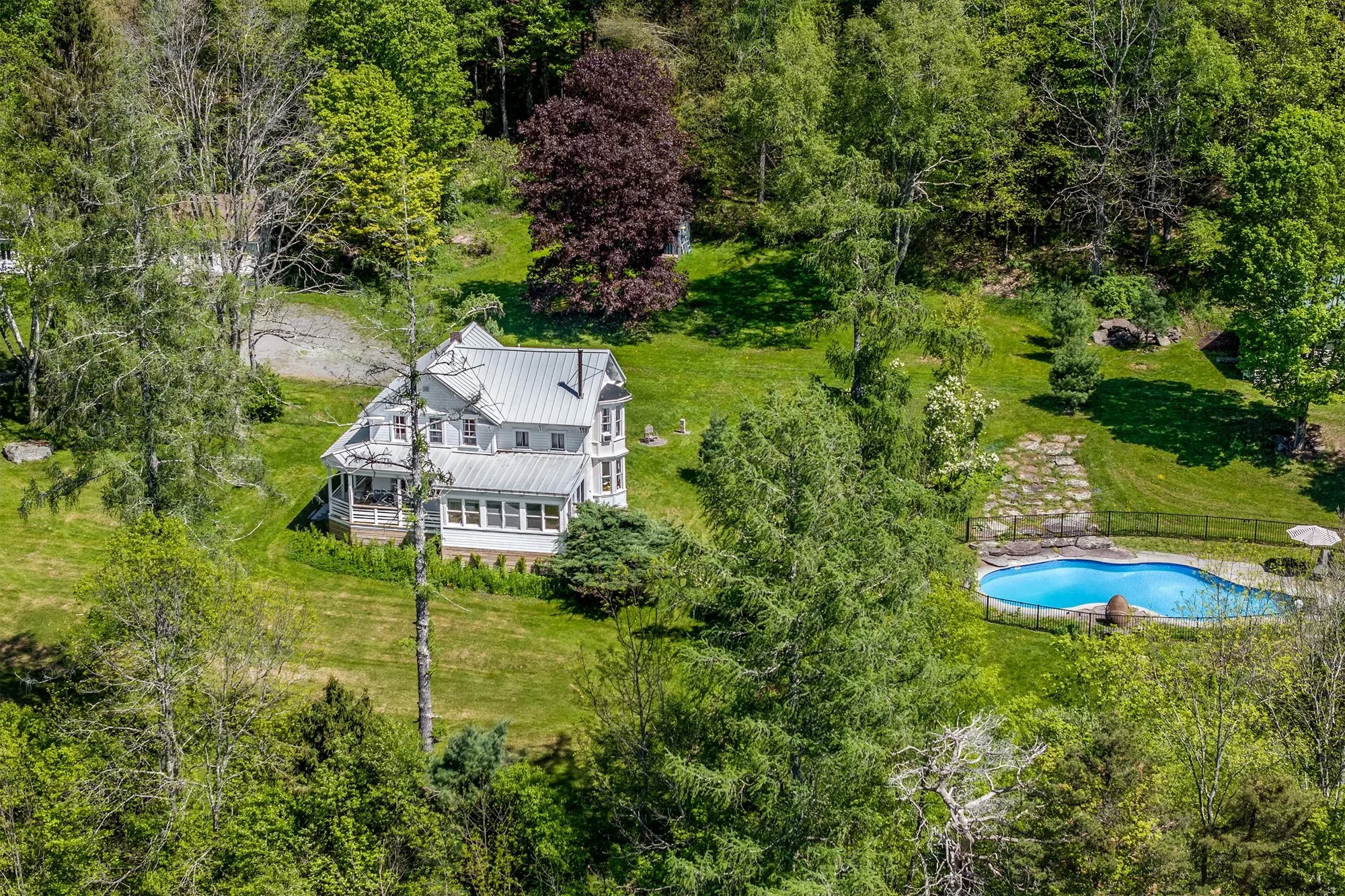 A white house with a gray metal roof situated in a lush, green backyard with tall trees and a swimming pool surrounded by a black fence.